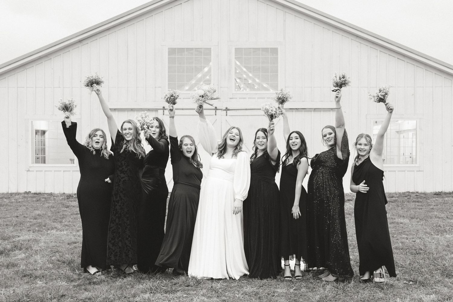 Wedding party in black dresses poses with bride in white against rustic barn backdrop.