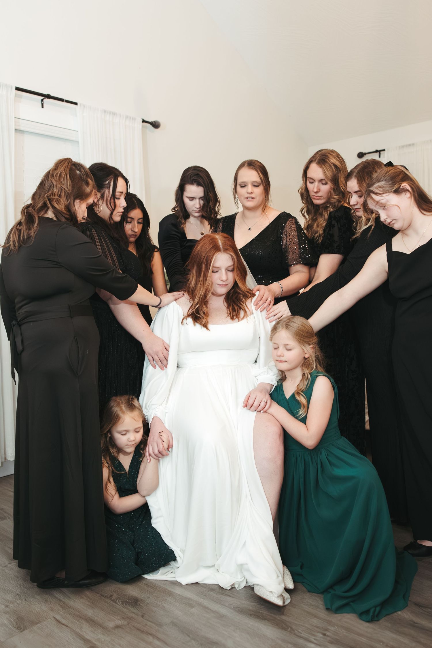 A bridal party in black dresses surrounds the bride in white, with two children in green dresses.