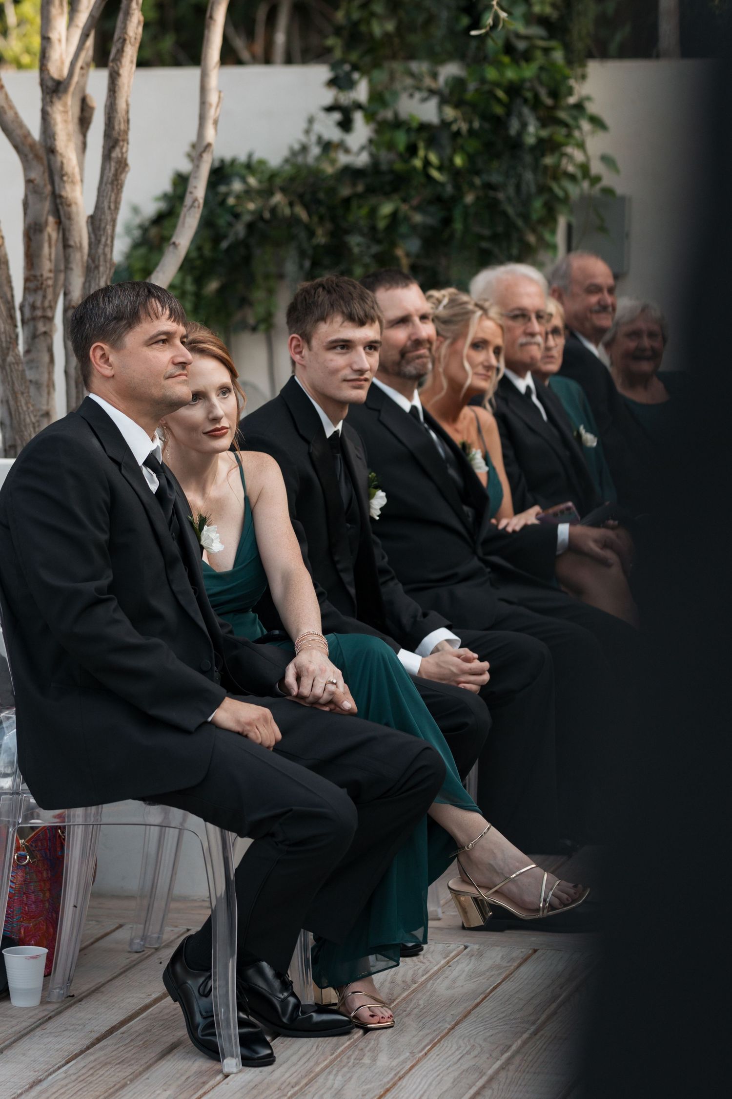 Wedding guests in formal attire seated on wooden benches during an indoor ceremony.