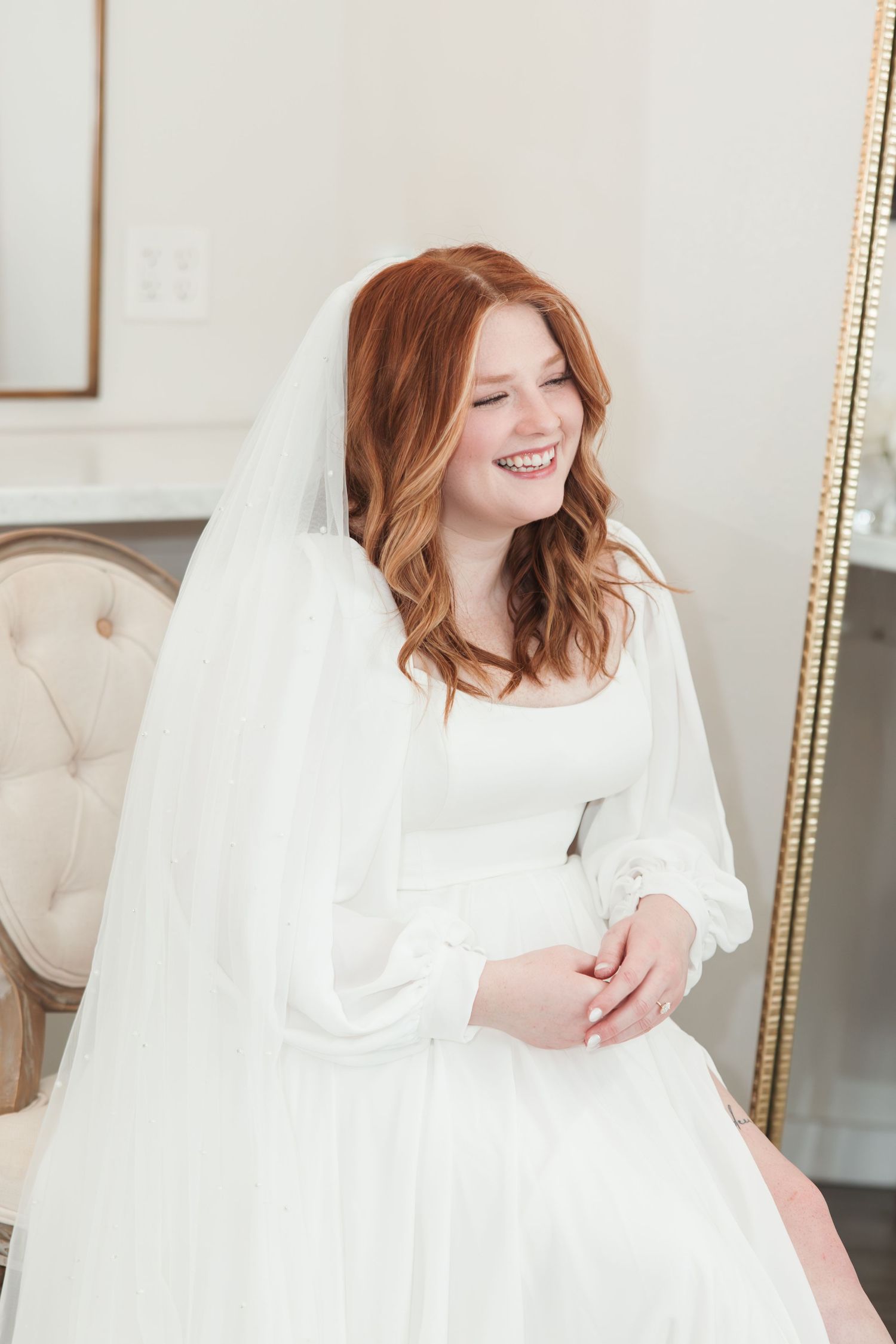A bride in a classic white wedding dress sits elegantly on a tufted chair with flowing red hair and a radiant smile.