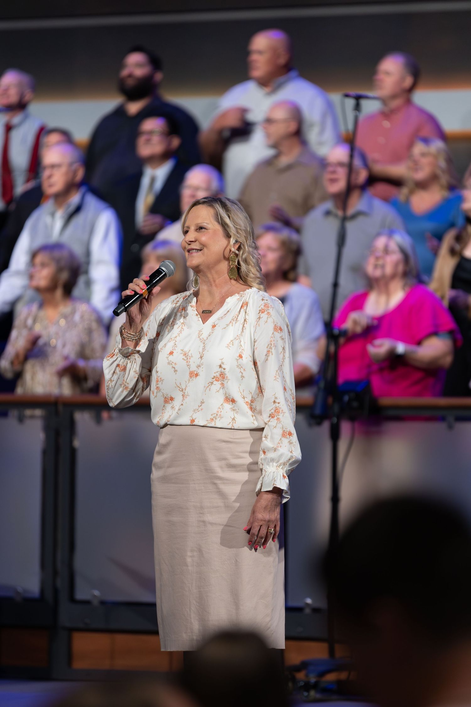 Singer performs with choir members visible in background on staged risers.