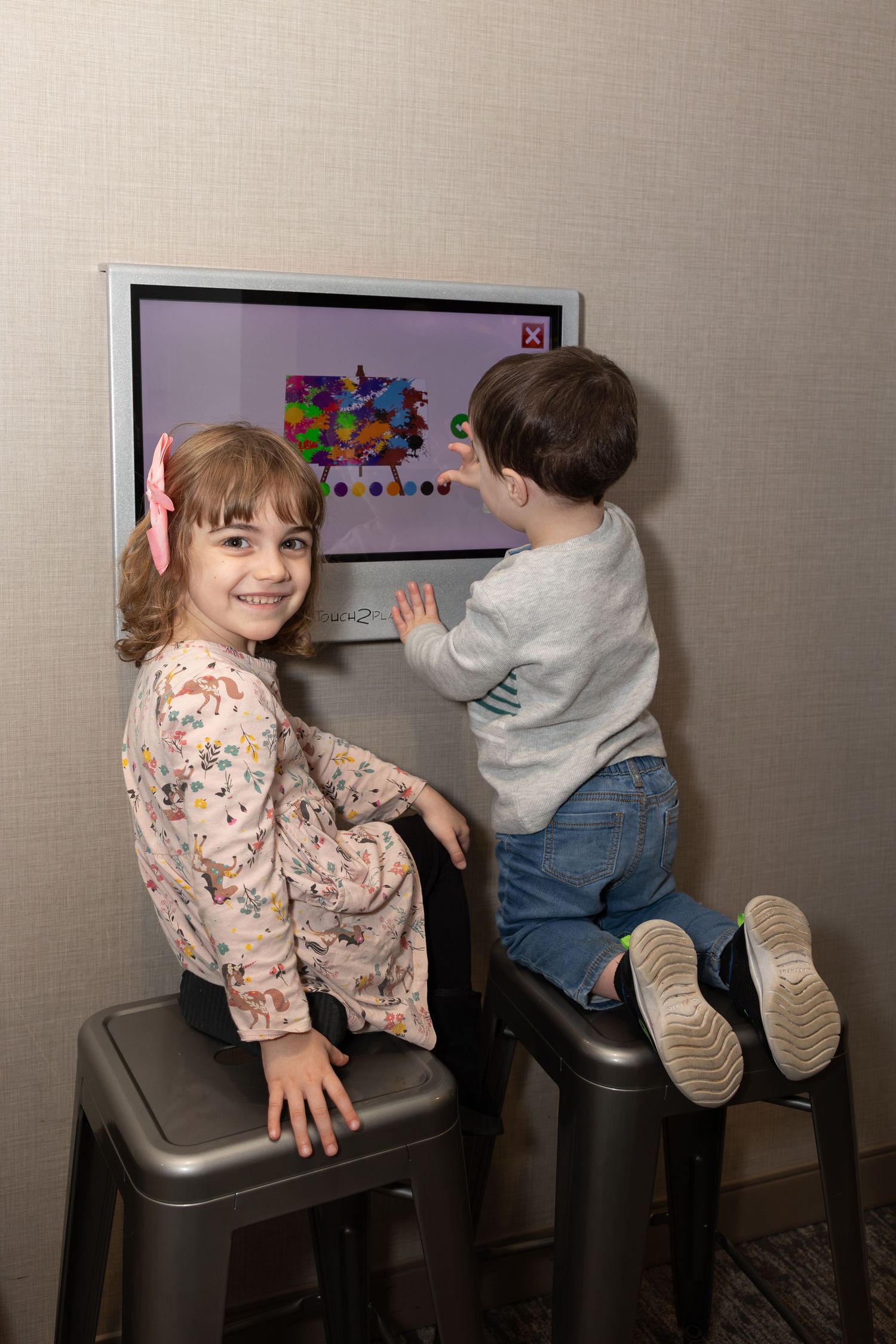 Two children interacting with a digital display screen while standing on stools.