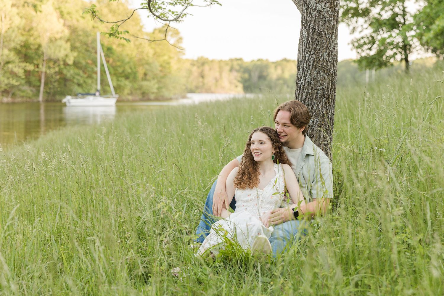 A couple sits embracing in tall grass near a lake with a sailboat in the background during golden hour.