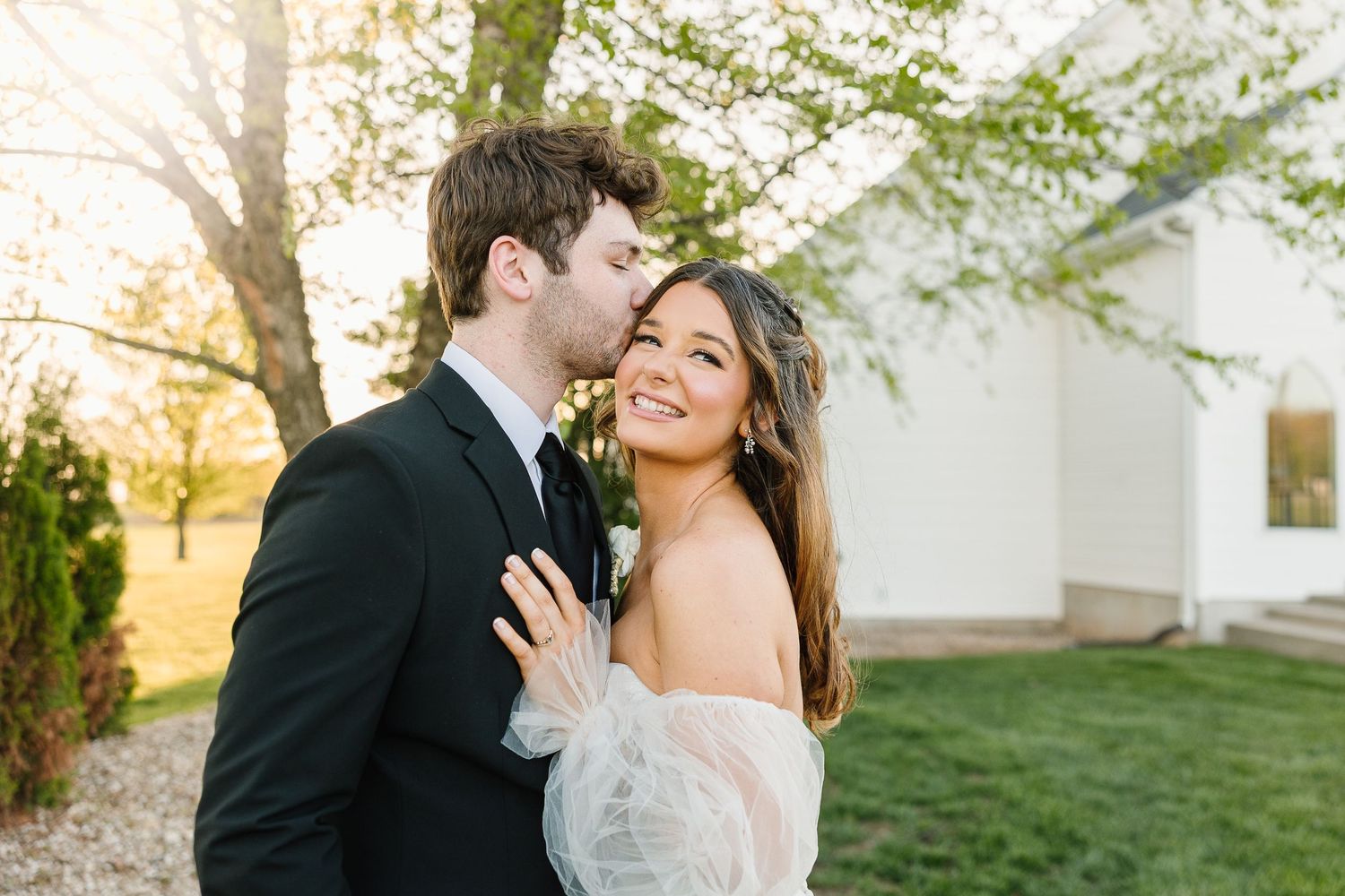 Romantic couple in formal attire share an intimate moment outdoors with a white building and trees in the background.