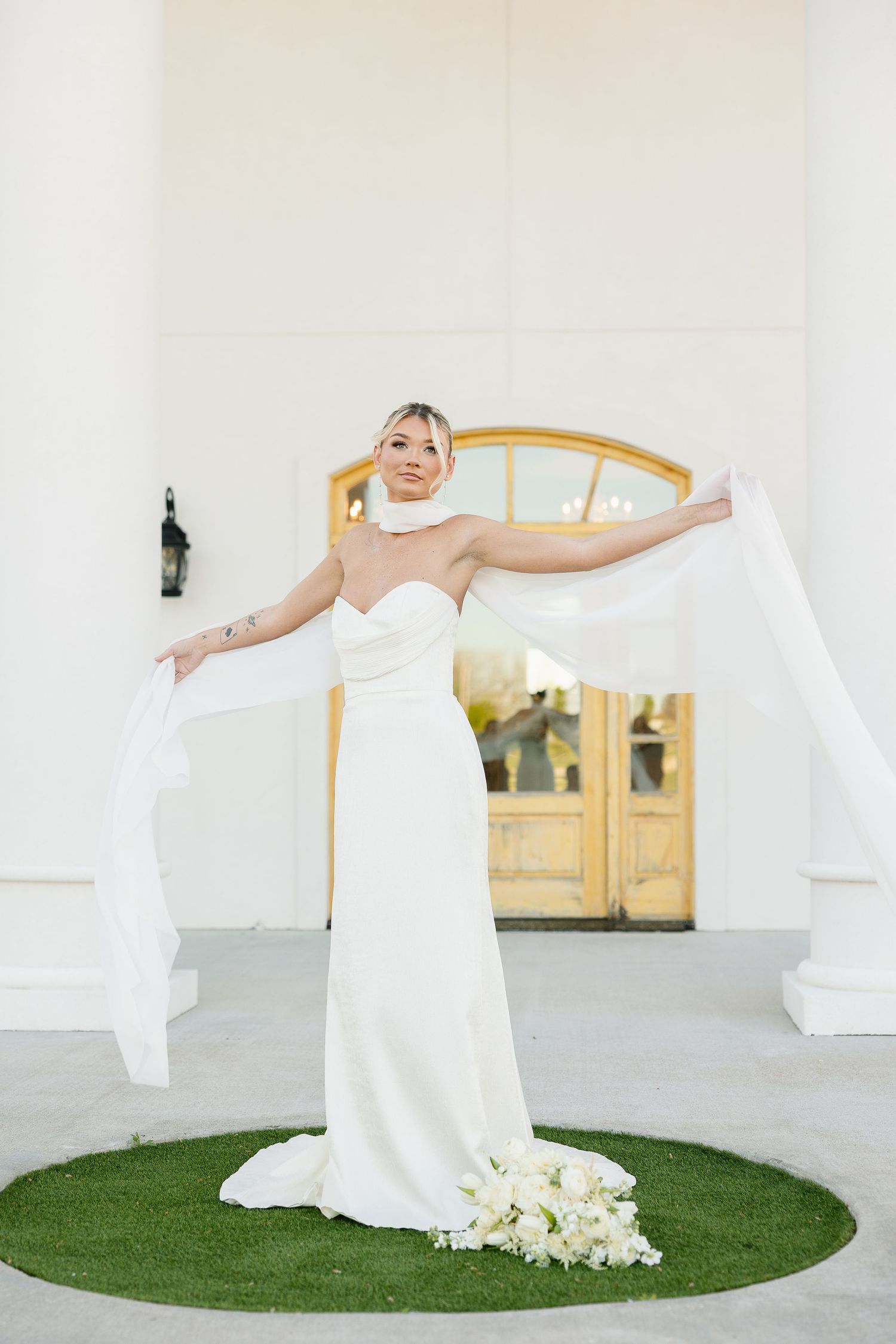 Bride in flowing white wedding dress twirls joyfully on circular grass platform against white building.