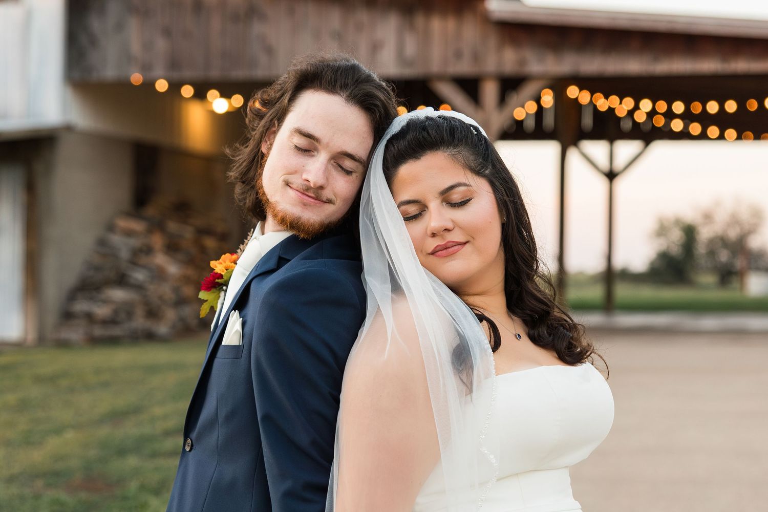 A romantic wedding couple embraces outdoors at sunset with string lights glowing in the background.