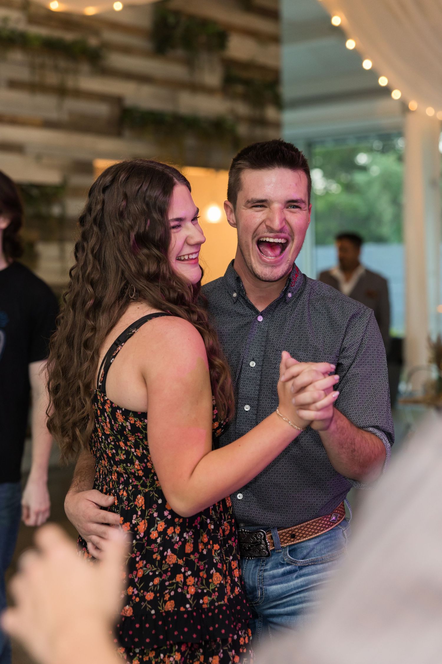 A joyful couple laughing and dancing together at a wedding reception with warm string lights in the background.