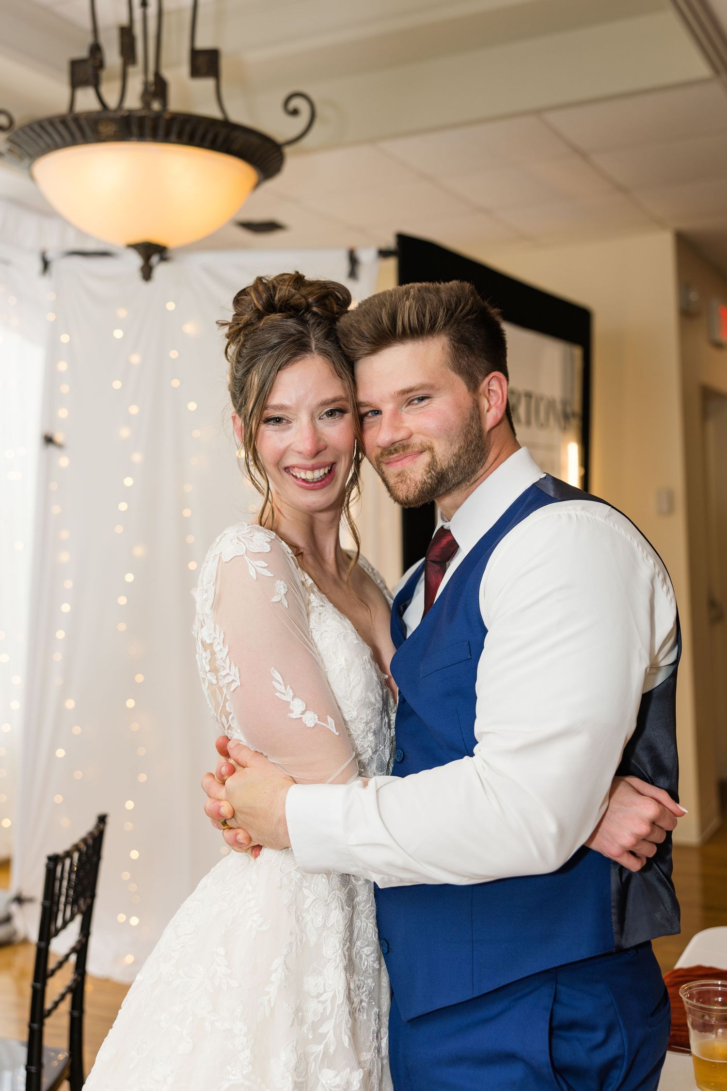 Bride and groom share first dance embrace smiling at wedding reception with string light backdrop.