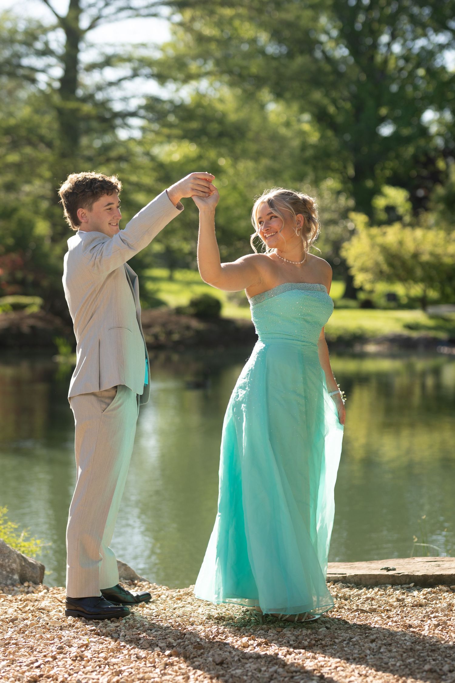 Young couple in prom attire dancing by a pond, girl in teal gown and boy in gray suit, surrounded by lush greenery.