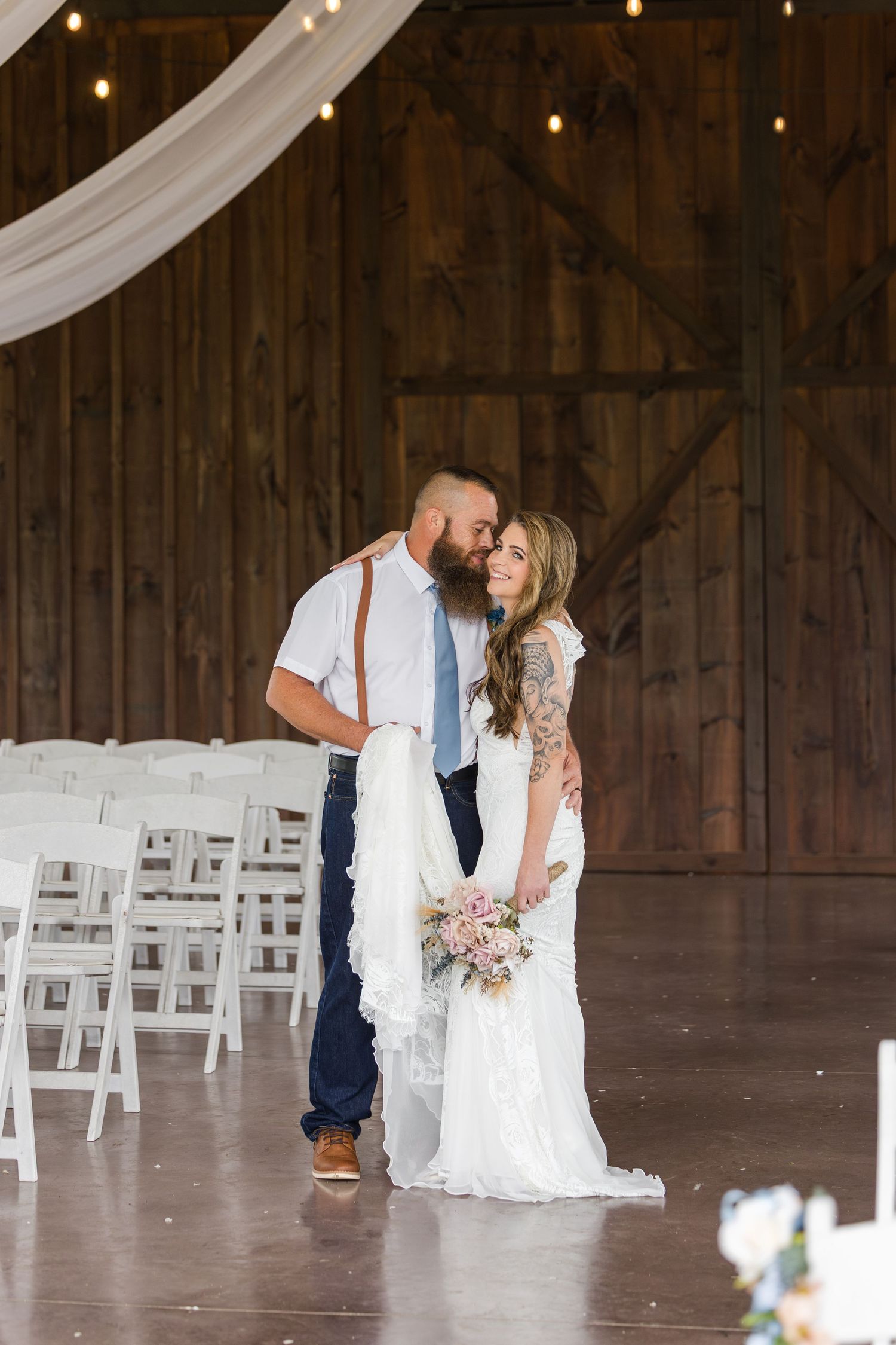 Bride and groom share a romantic moment in a rustic barn wedding venue with white chairs and string lights.