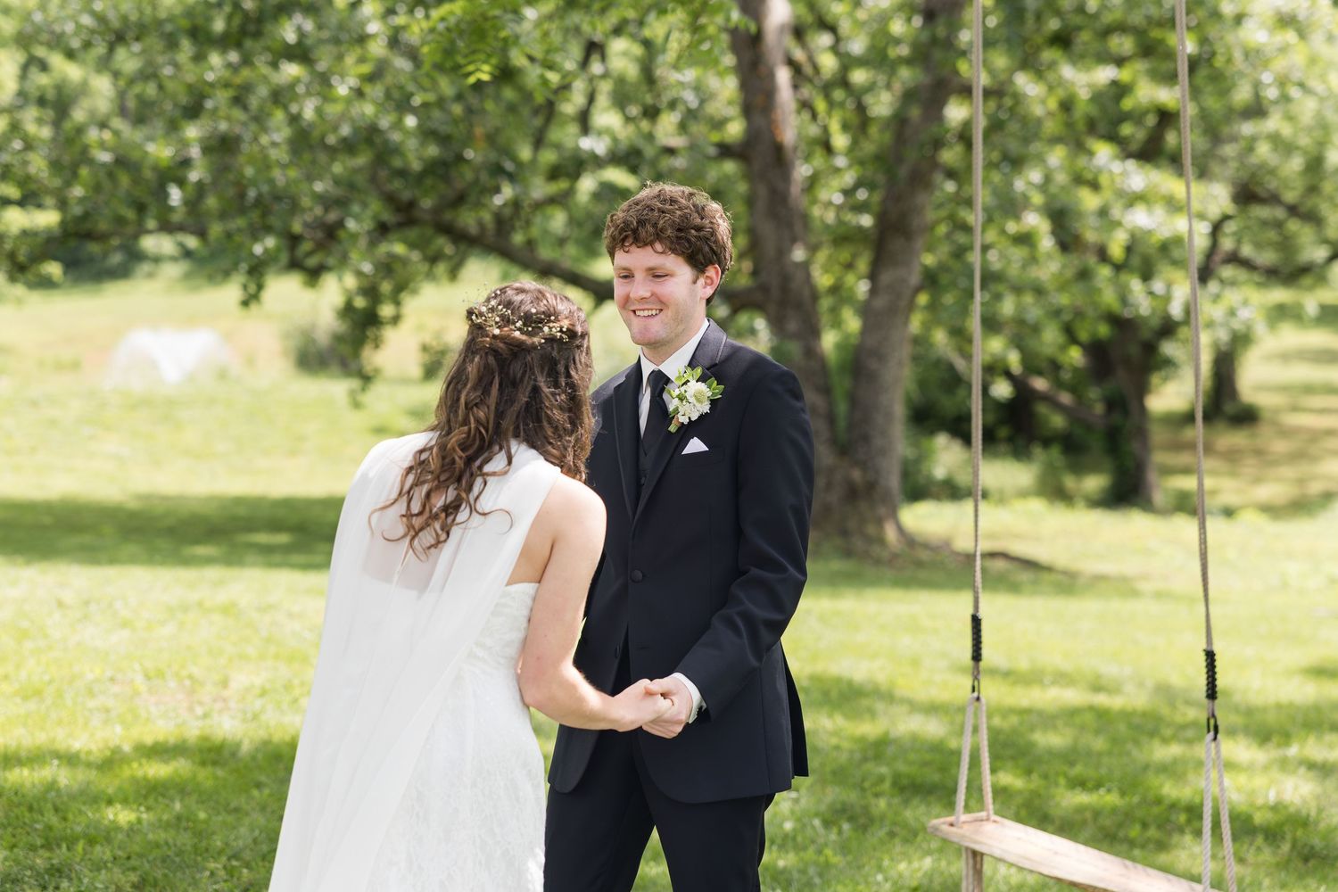 Bride and groom holding hands during outdoor wedding ceremony on green lawn with swing and trees in background.