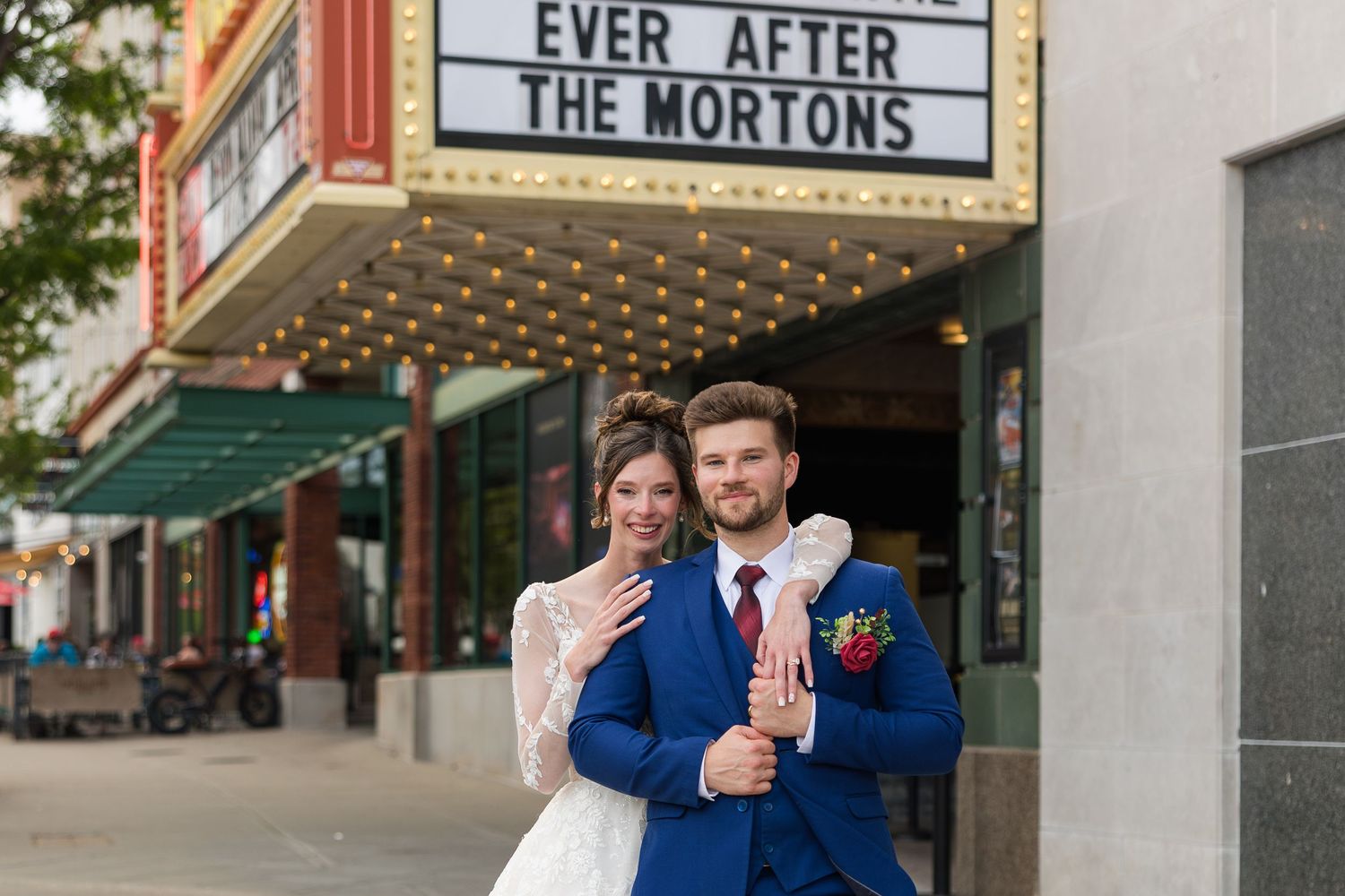 Newlyweds pose outside a theater marquee reading Ever After The Mortons, bride showing ring in wedding attire.
