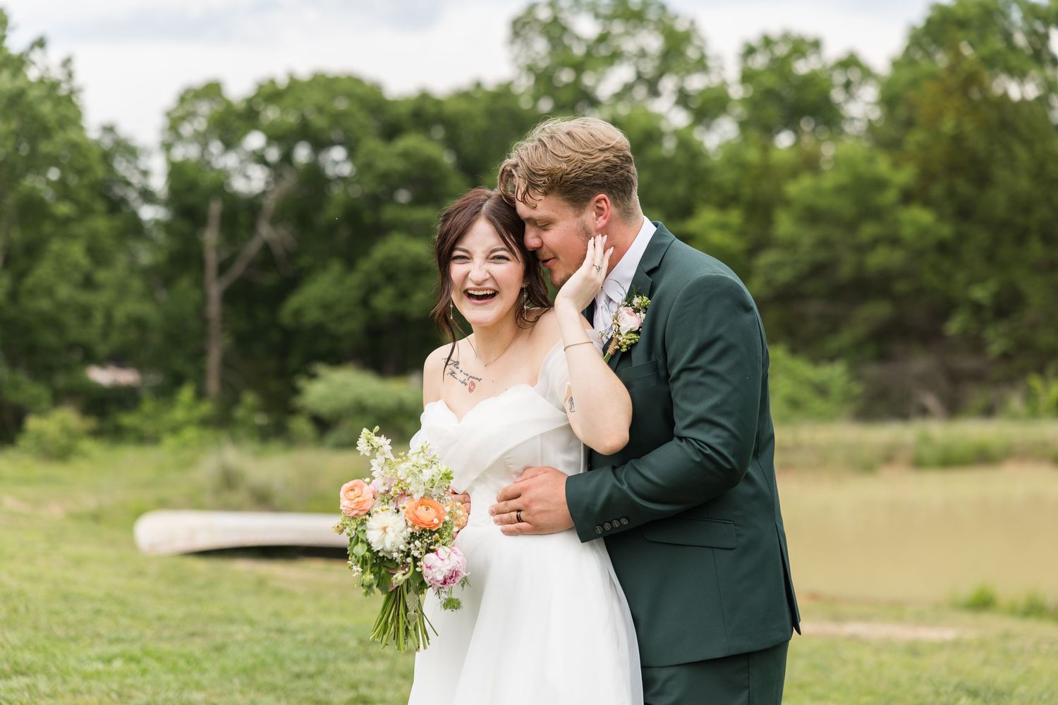 Joyful bride and groom share a laugh outdoors, she holds a colorful bouquet, he wears a dark green suit.