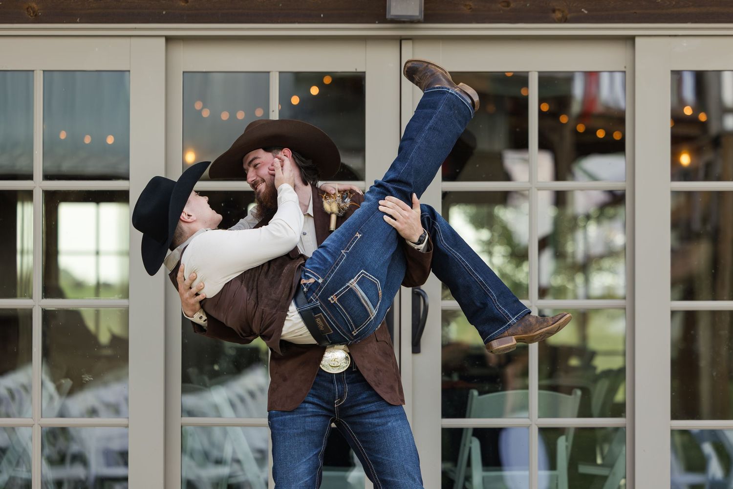 Cowboy groom lifts his bride in jeans outdoors near glass doors at a rustic western-themed wedding celebration.
