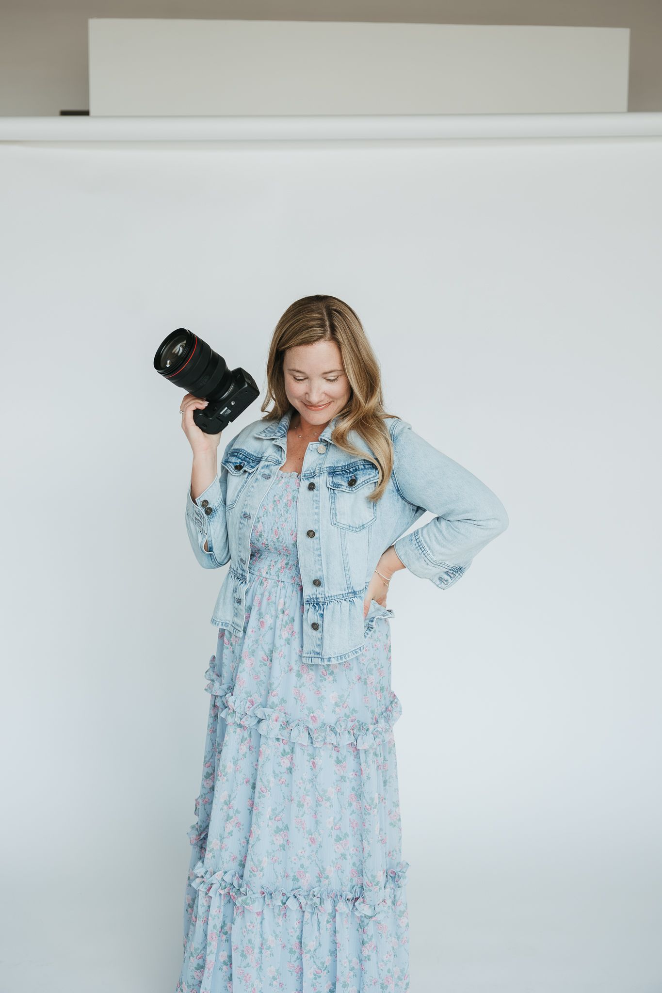 A photographer in a flowy light blue floral maxi dress holds a professional camera against a white background.
