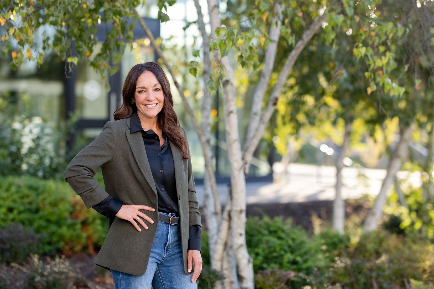 Professional woman in blazer and jeans poses confidently outdoors against natural birch tree backdrop.