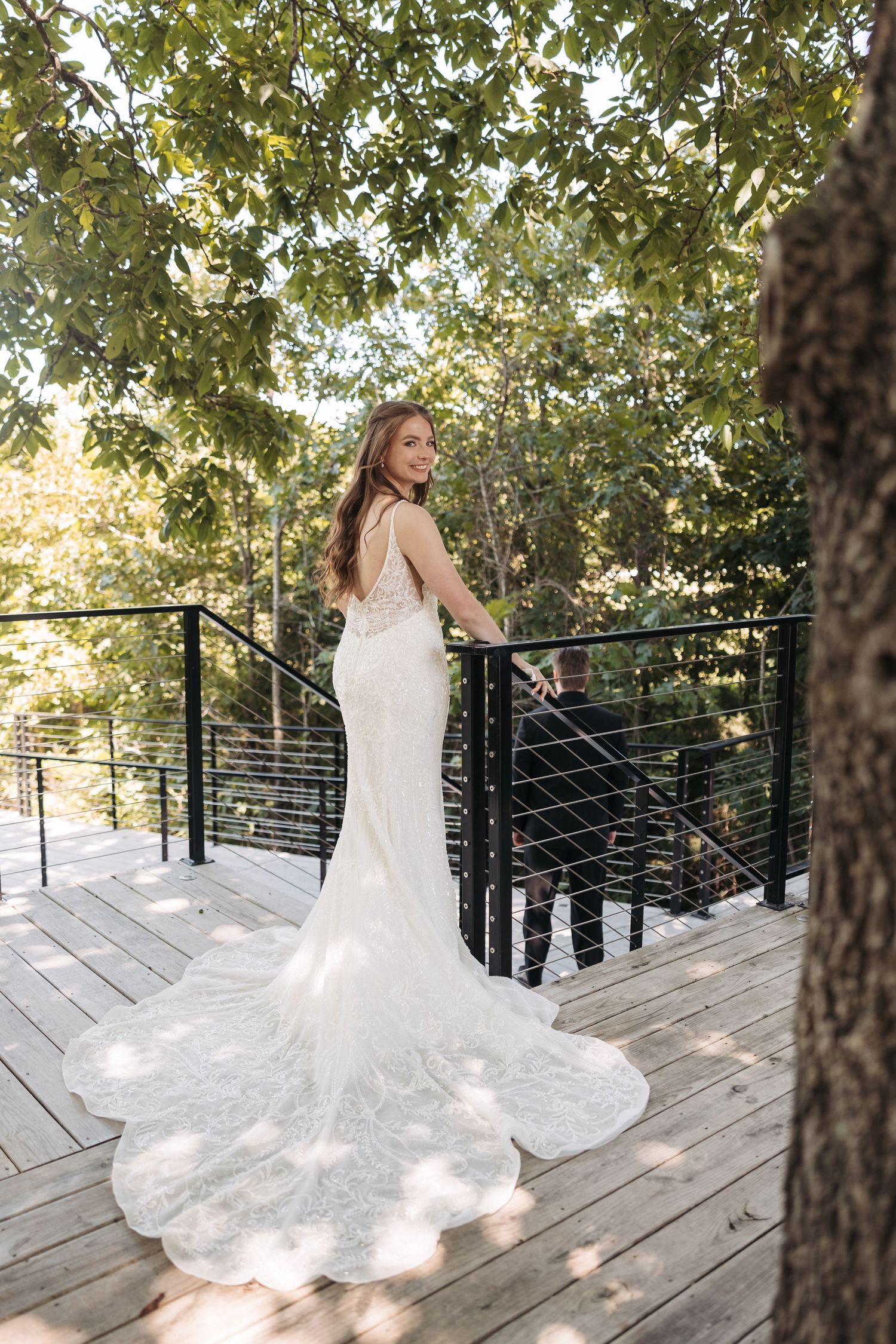 Bride in fitted lace gown with long train poses on wooden deck under dappled sunlight.