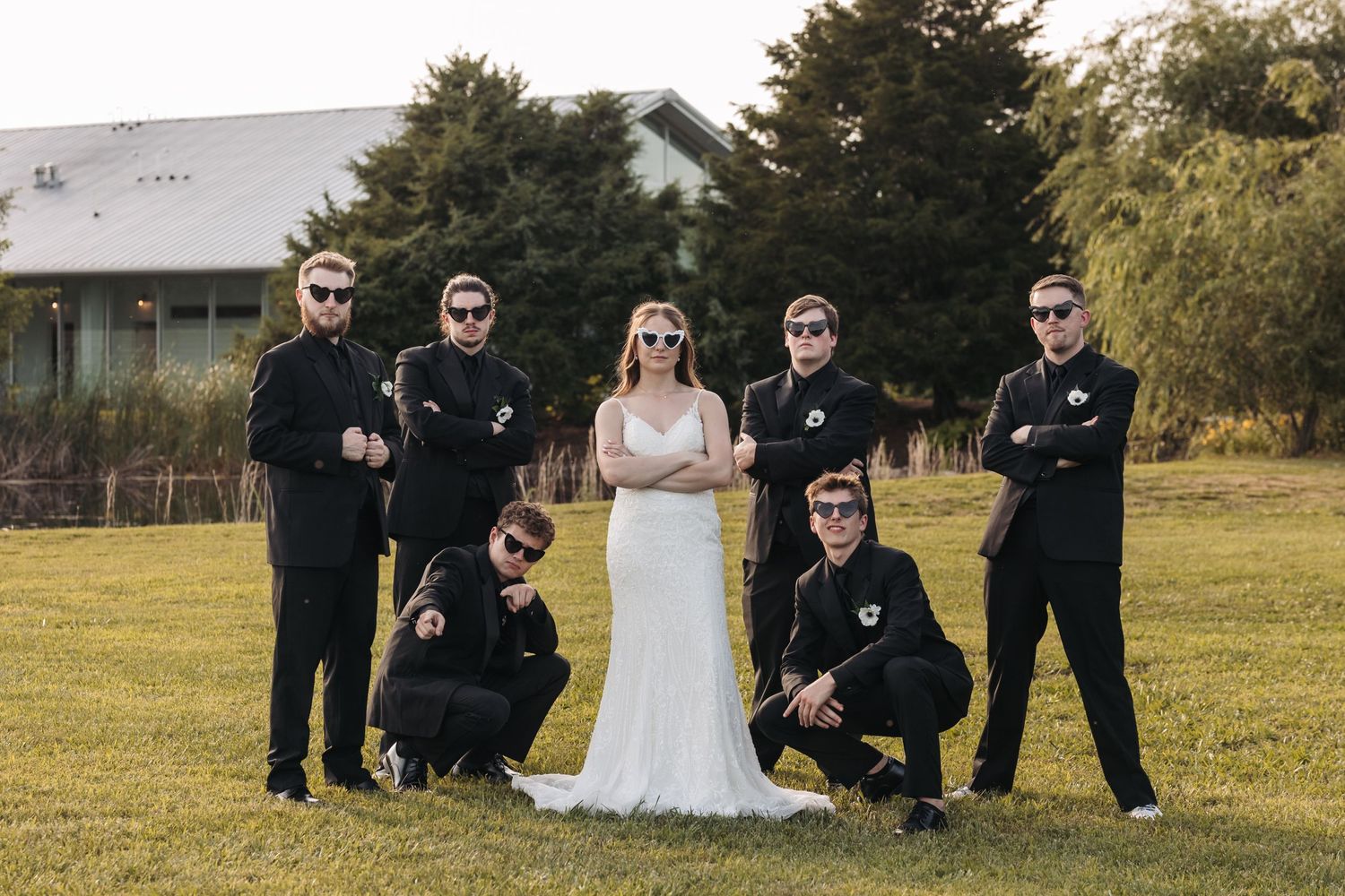 Wedding party poses in casual black attire with bride in white dress on grassy field.