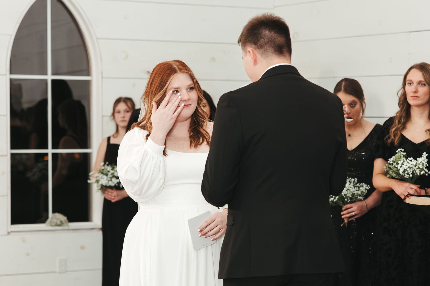 An emotional moment during a wedding ceremony with bridesmaids in black dresses watching in background.