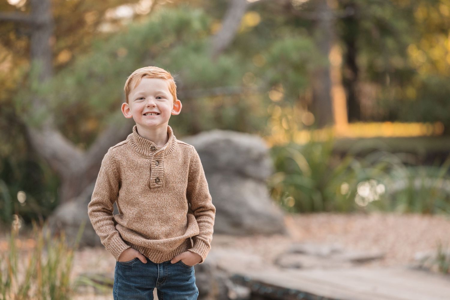 A child in a brown sweater smiles outdoors with autumn trees in background.