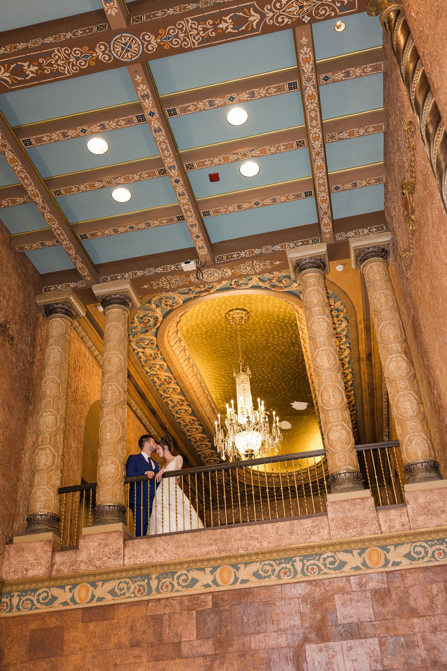 Bride and groom share a romantic kiss on a balcony inside an ornate golden wedding venue with elegant columns.