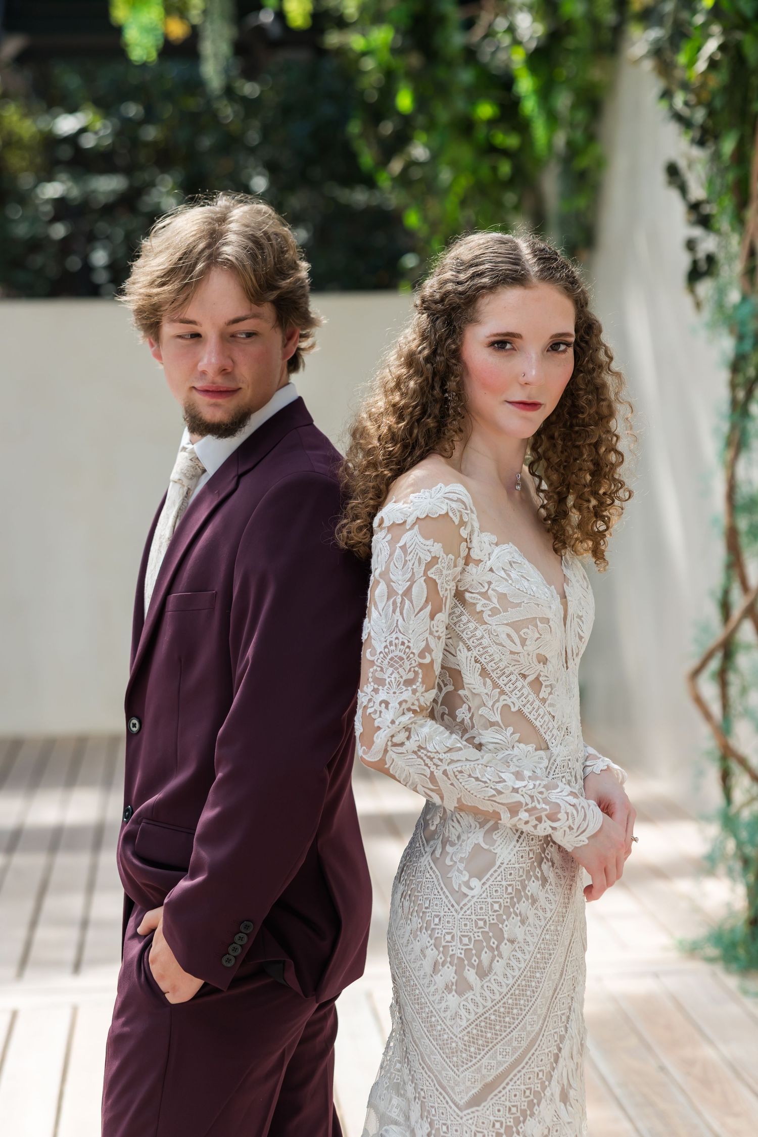 Bride in lace gown and groom in burgundy suit pose back-to-back in an outdoor garden setting.