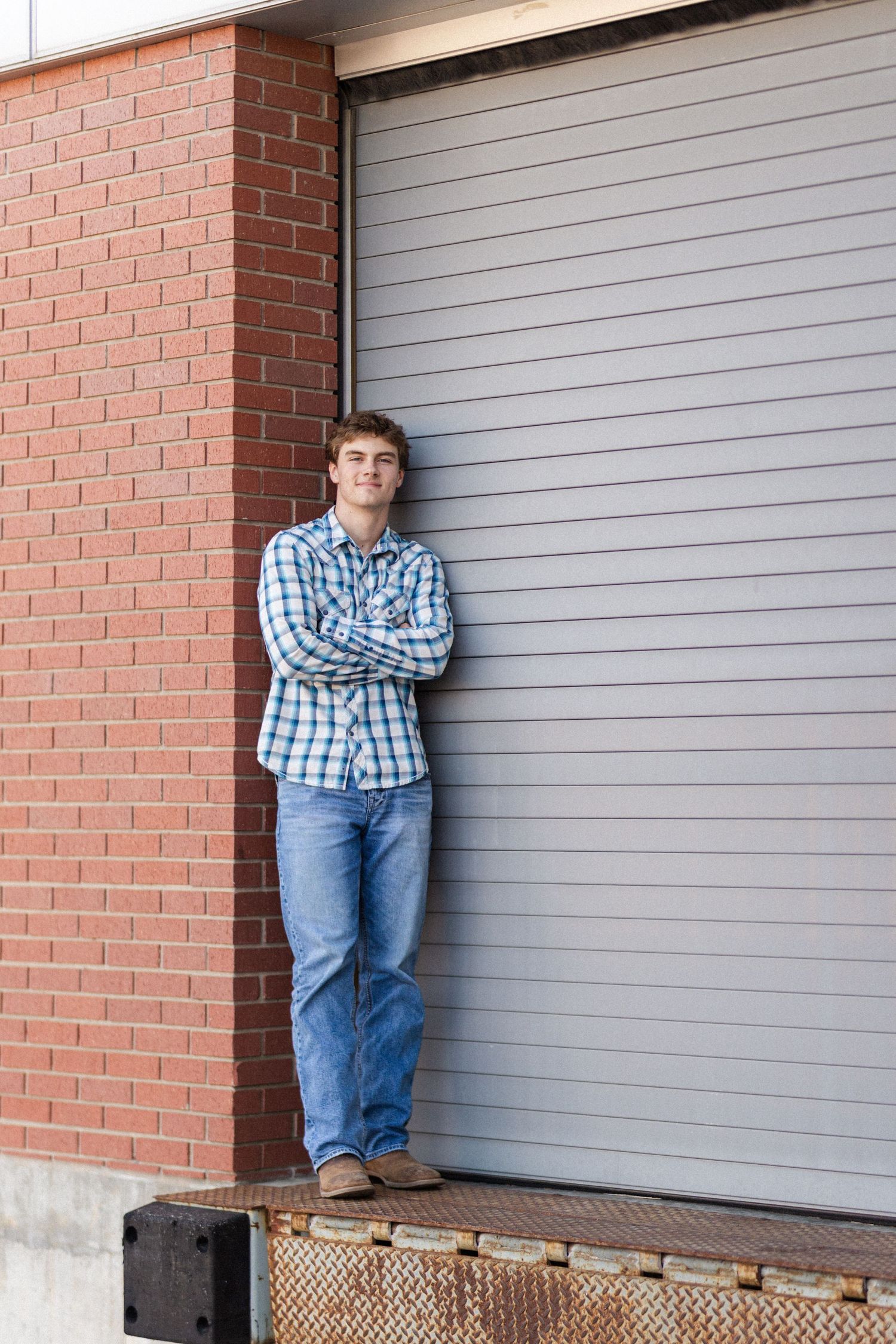 Person in plaid shirt and jeans leaning against brick wall by loading dock door.