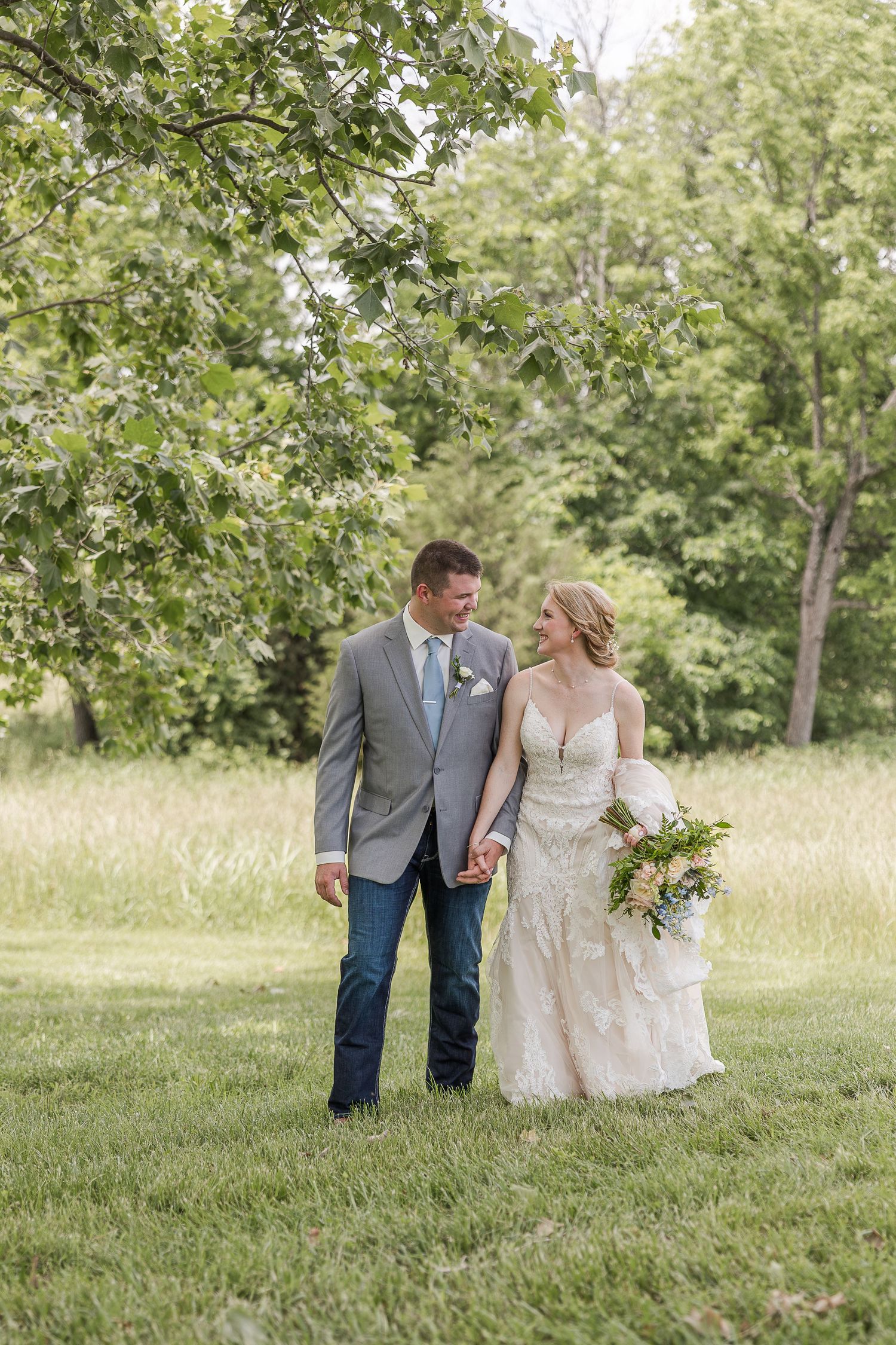 Newlywed couple holding hands in outdoor field with trees, groom in gray jacket and jeans, bride in lace gown with bouquet.