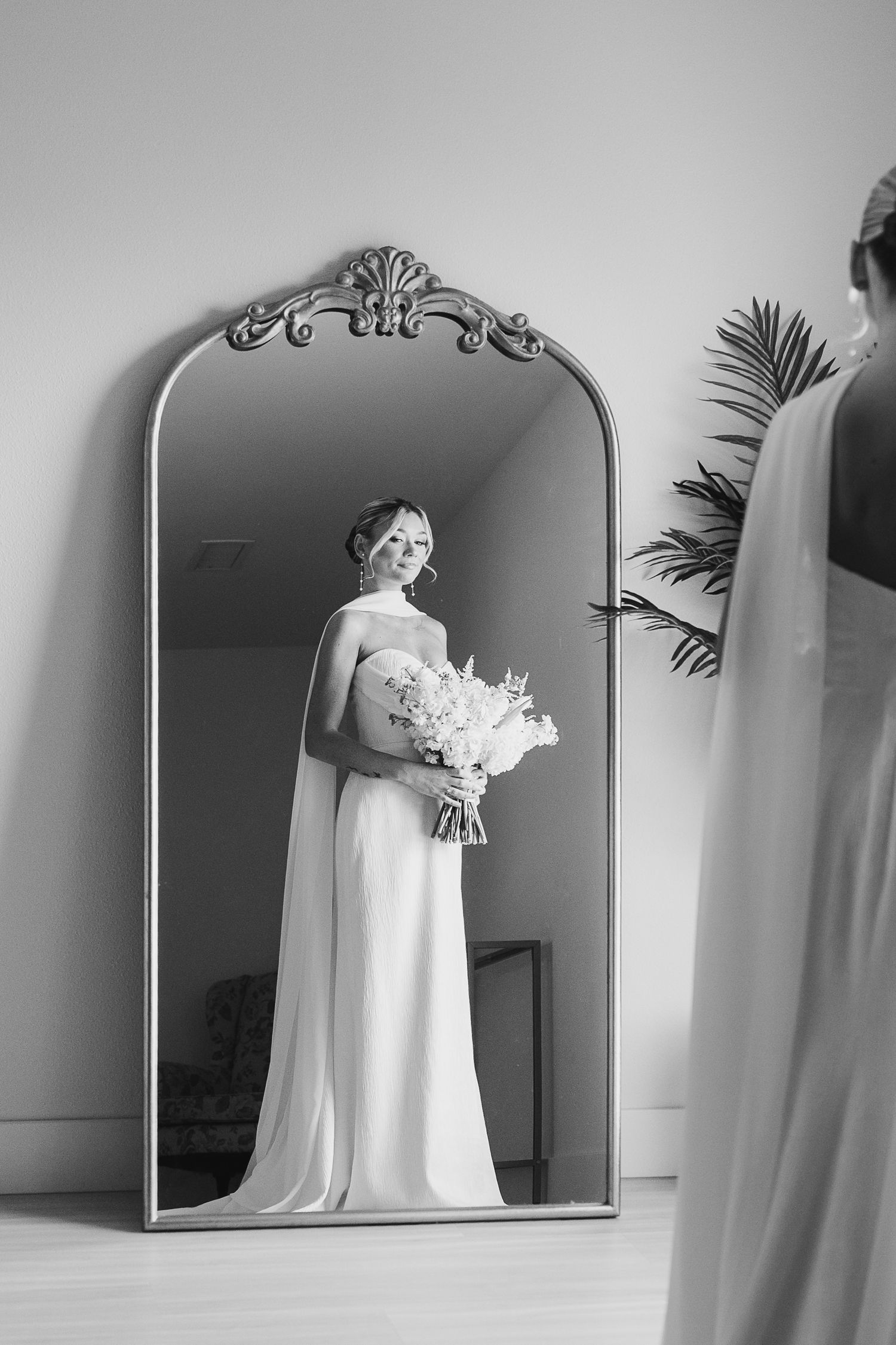 Elegant black and white reflection in ornate mirror shows bride in flowing dress holding white floral bouquet.