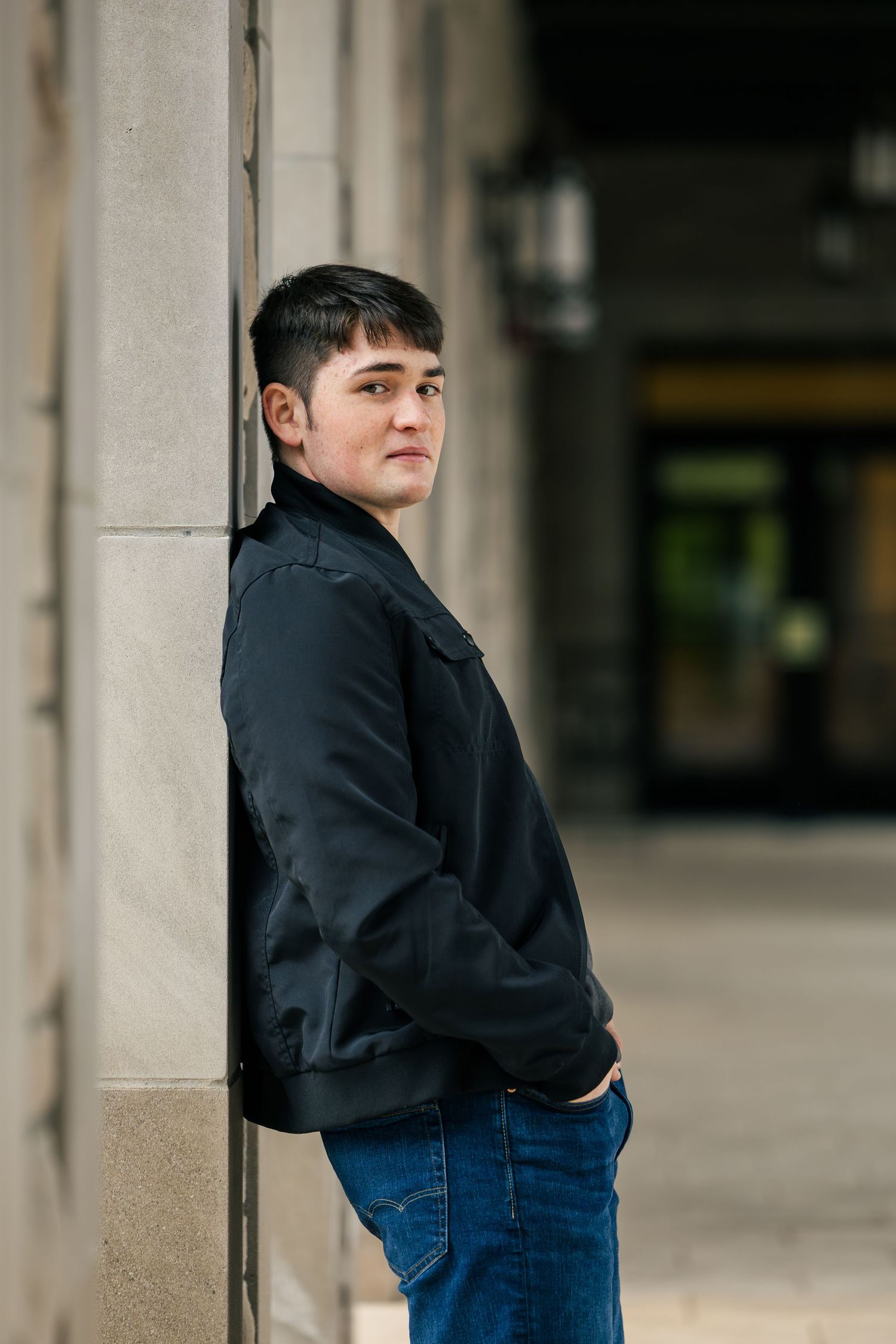 A person in a black jacket and blue jeans leans against a stone column in an urban setting.