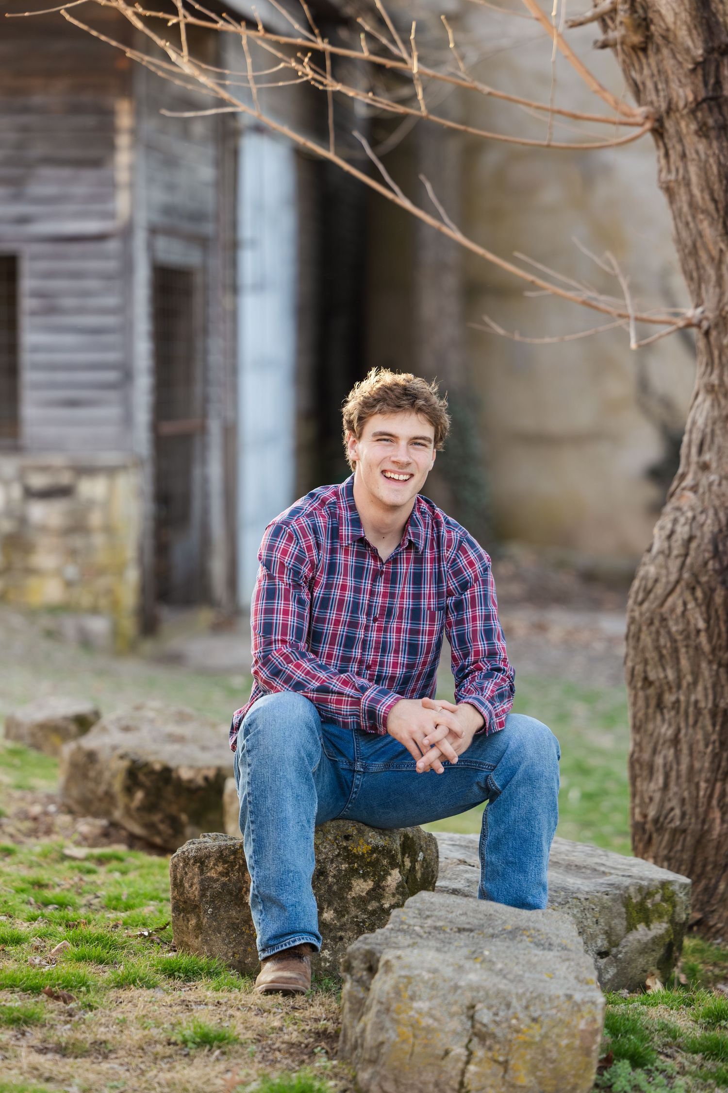 Casual portrait of person in plaid shirt sitting on rocks by rustic building.