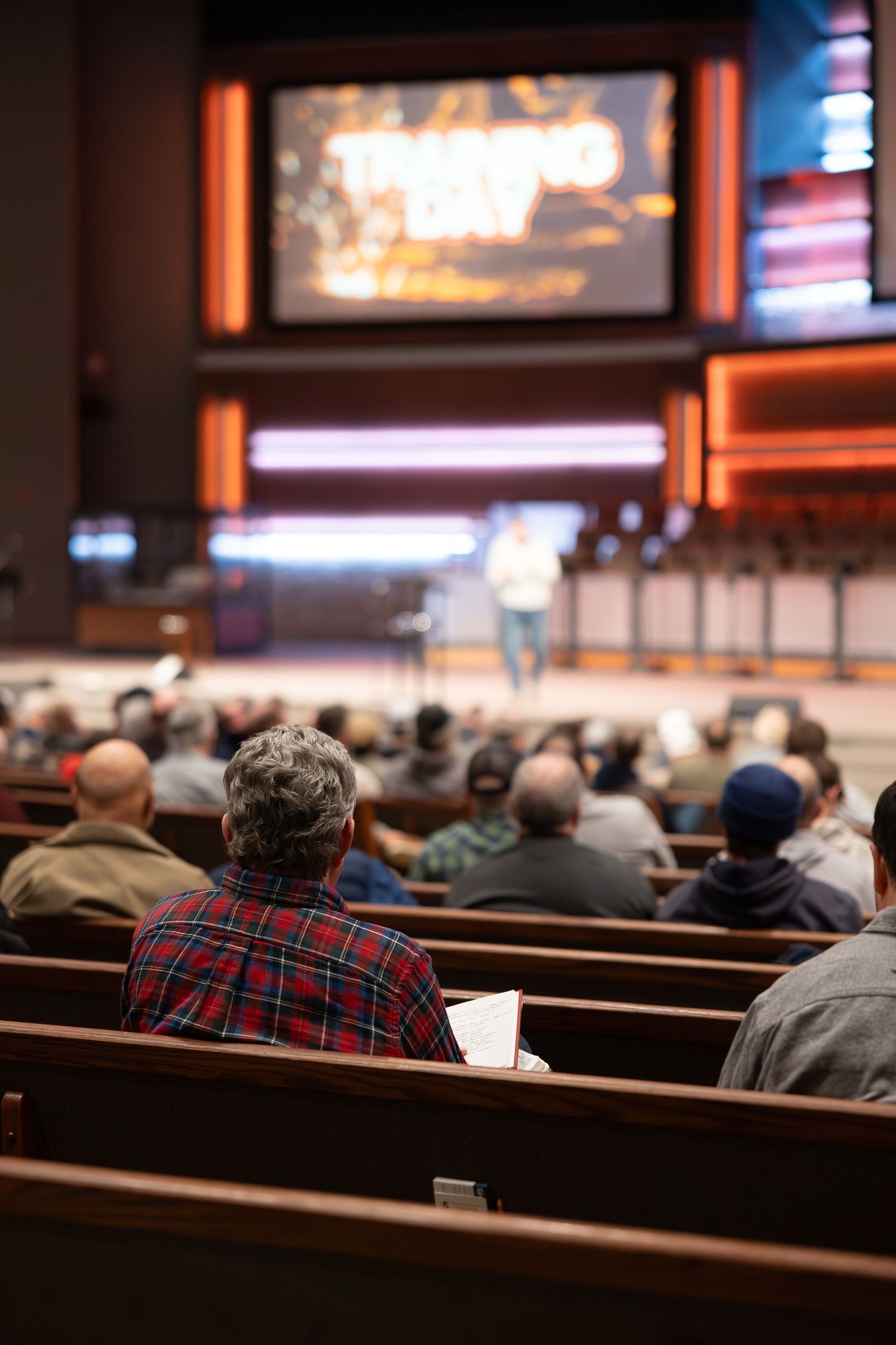 Audience members seated in wooden pews watch a large screen during a church service or presentation.