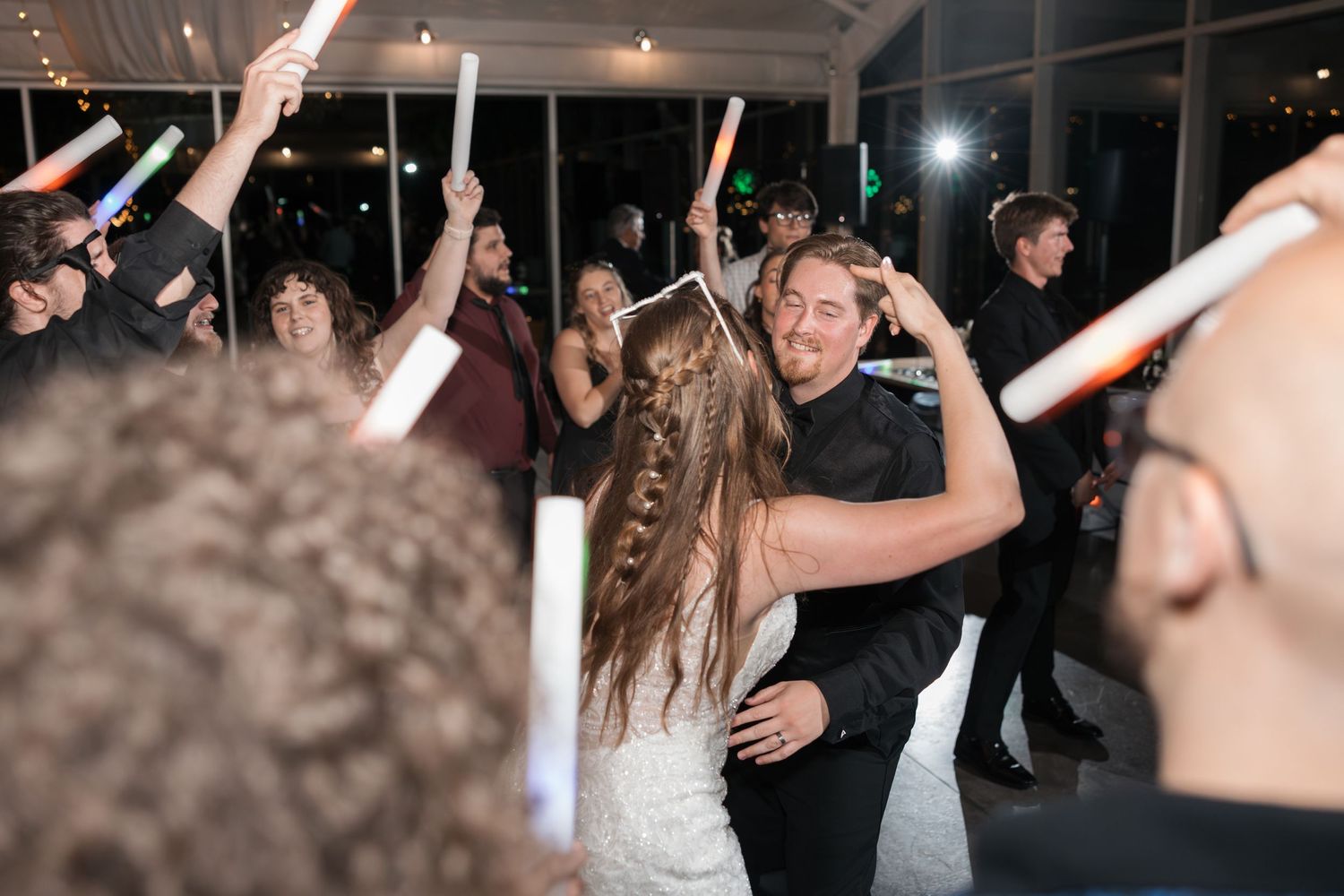 Wedding celebration shows dancing guests with light-up wands under indoor string lights.