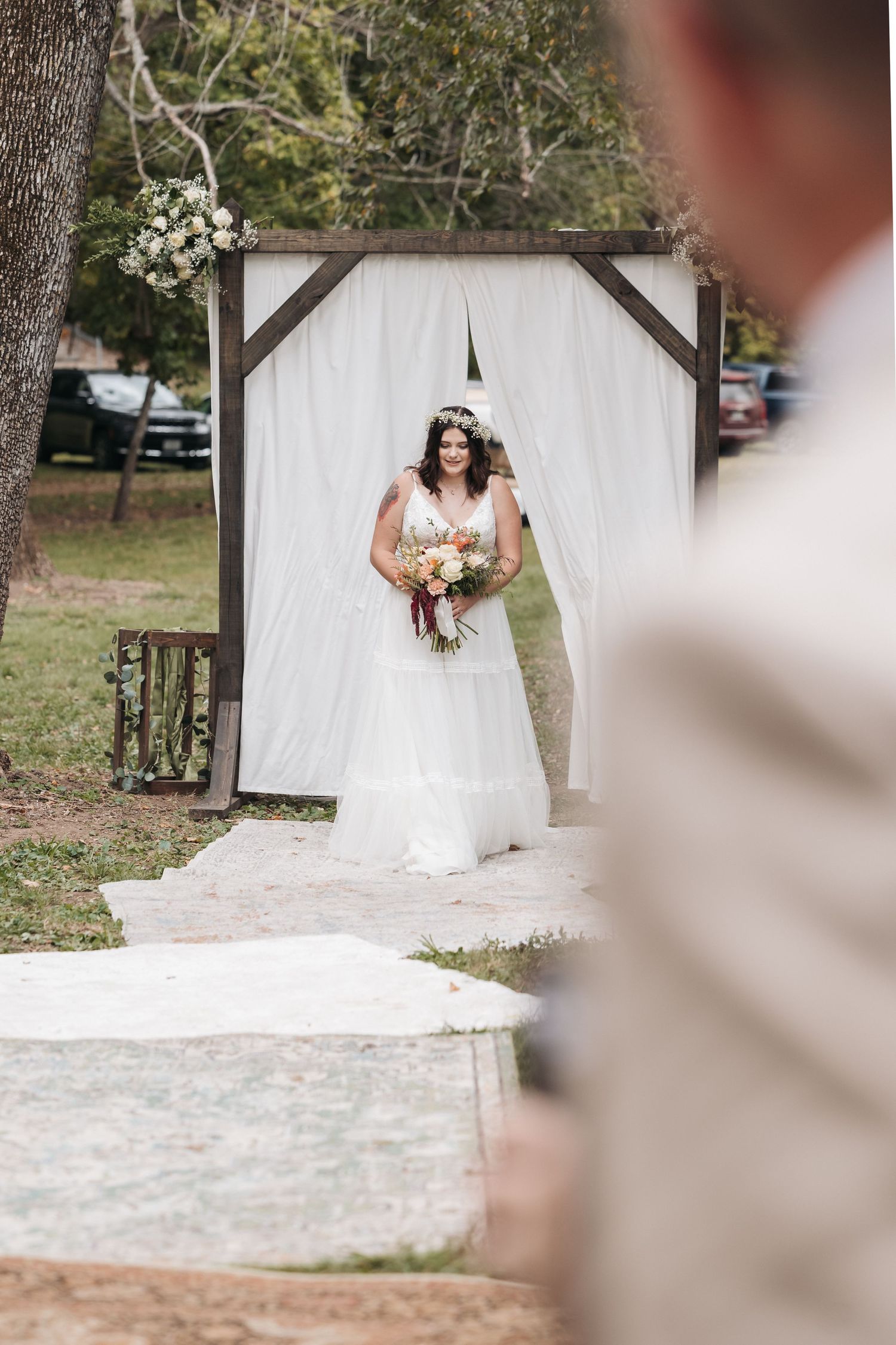 Wedding processional moment with white fabric backdrop and rustic wooden arch in outdoor setting.