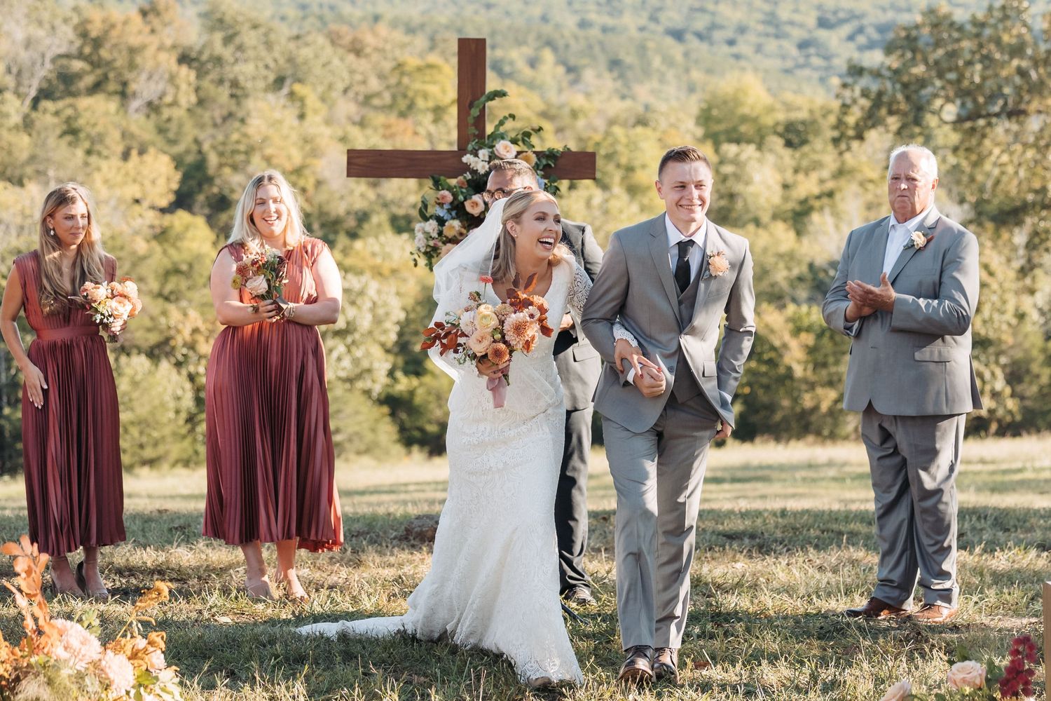 A wedding party stands near a wooden cross during an outdoor autumn ceremony.