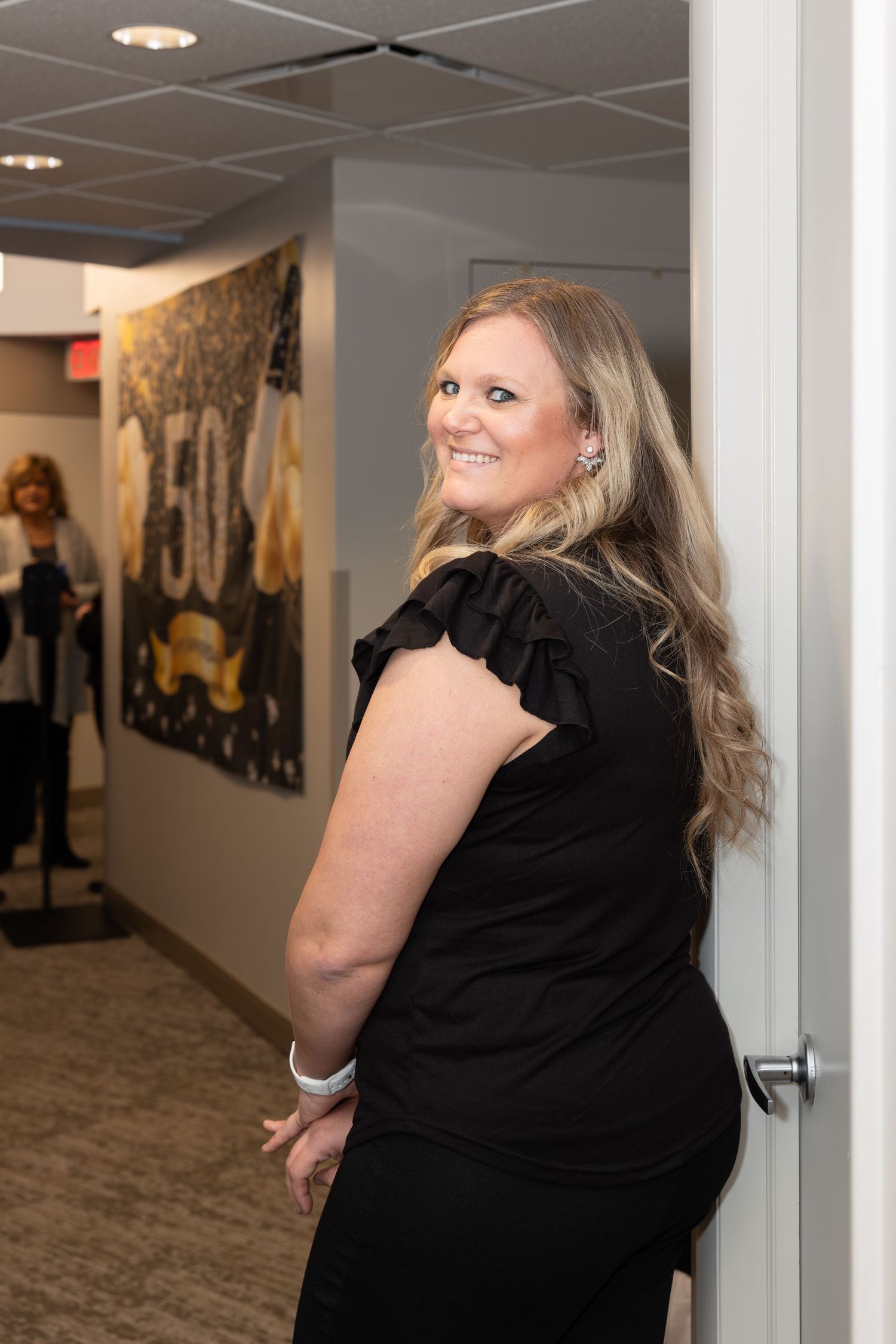 Staff member in black attire smiling over shoulder in office hallway.