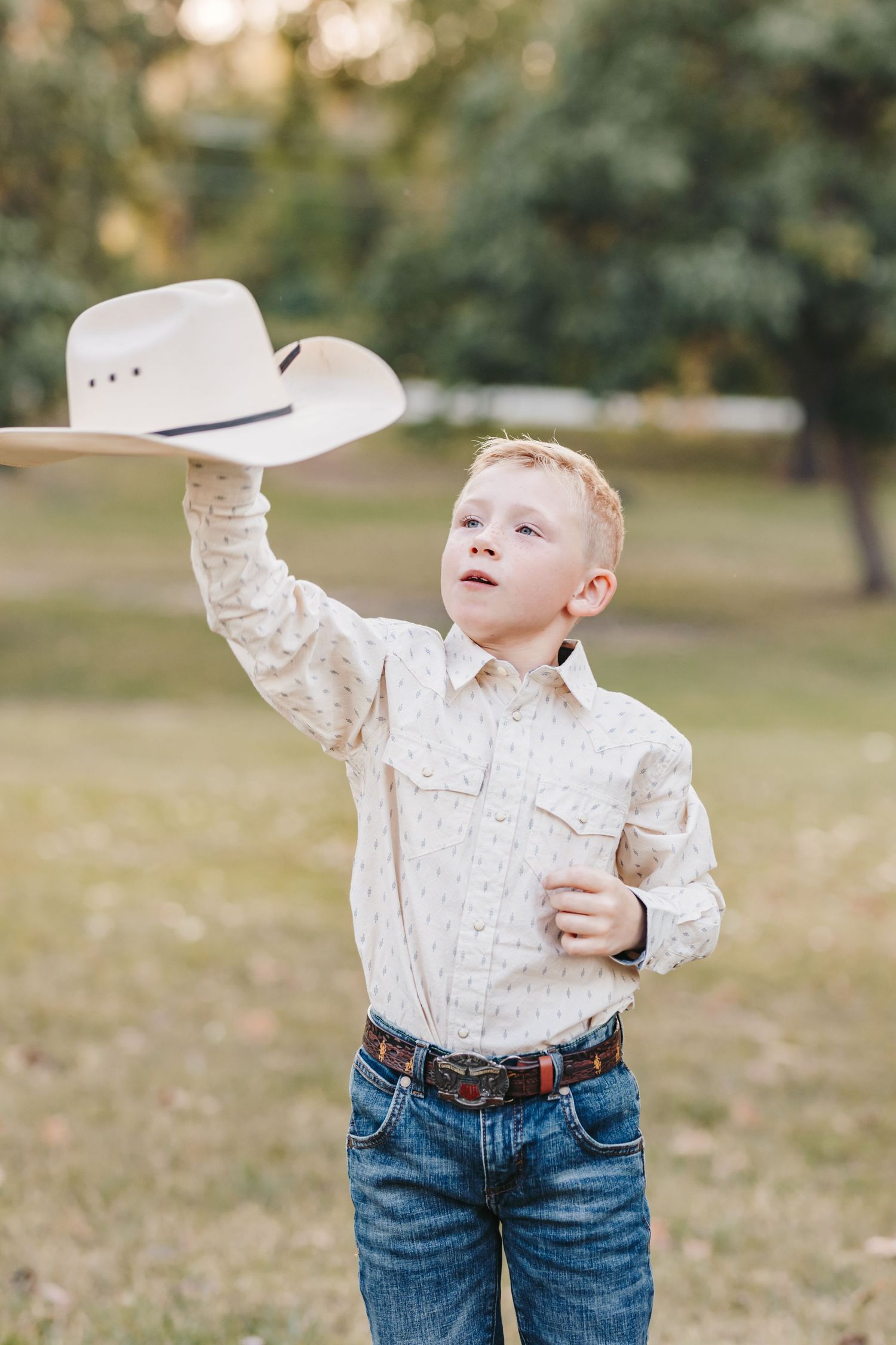 Young child in western wear holds up a white cowboy hat while standing outdoors on grass.