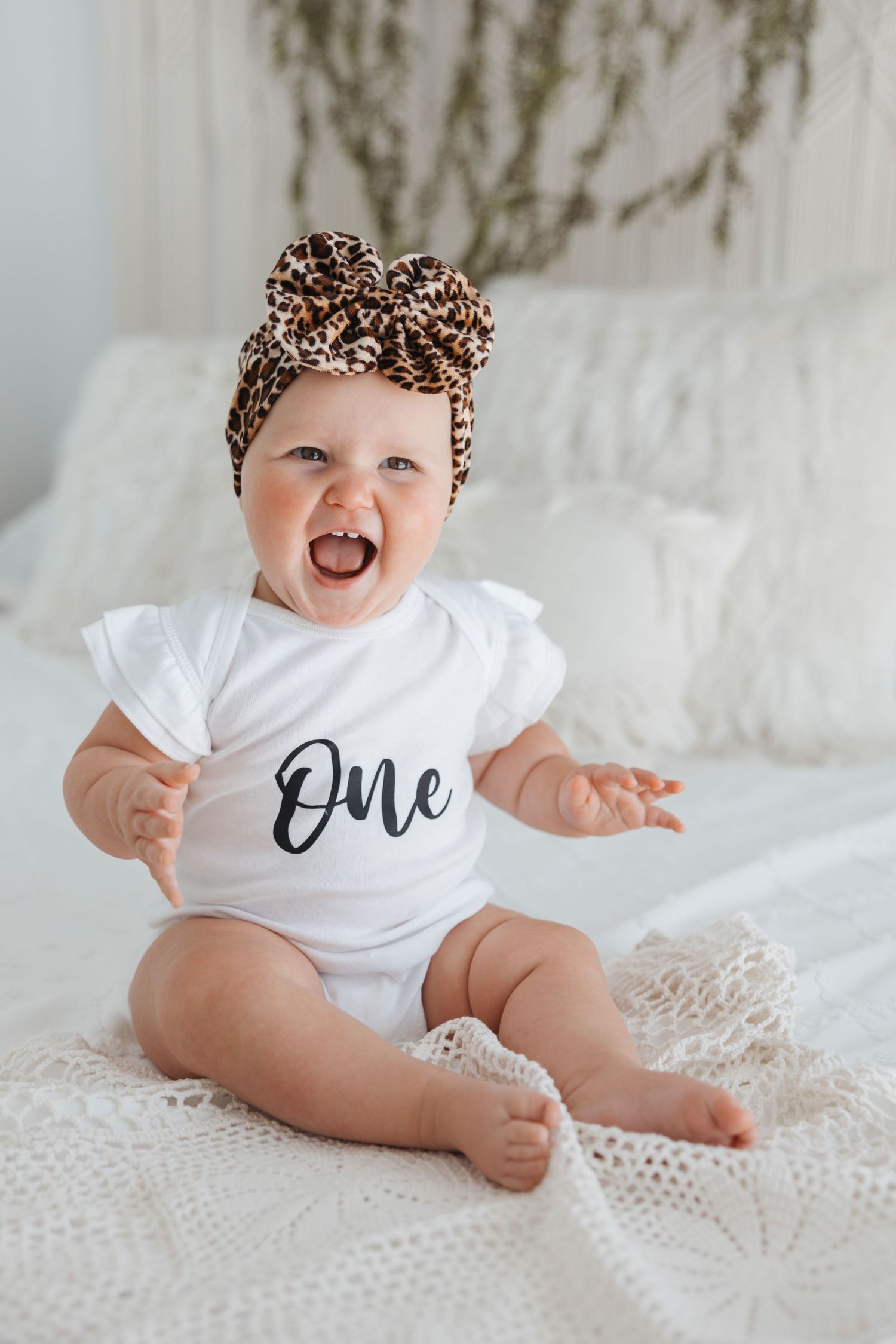 A baby wearing a white onesie with 'One' text and a leopard print headwrap sits on white bedding.