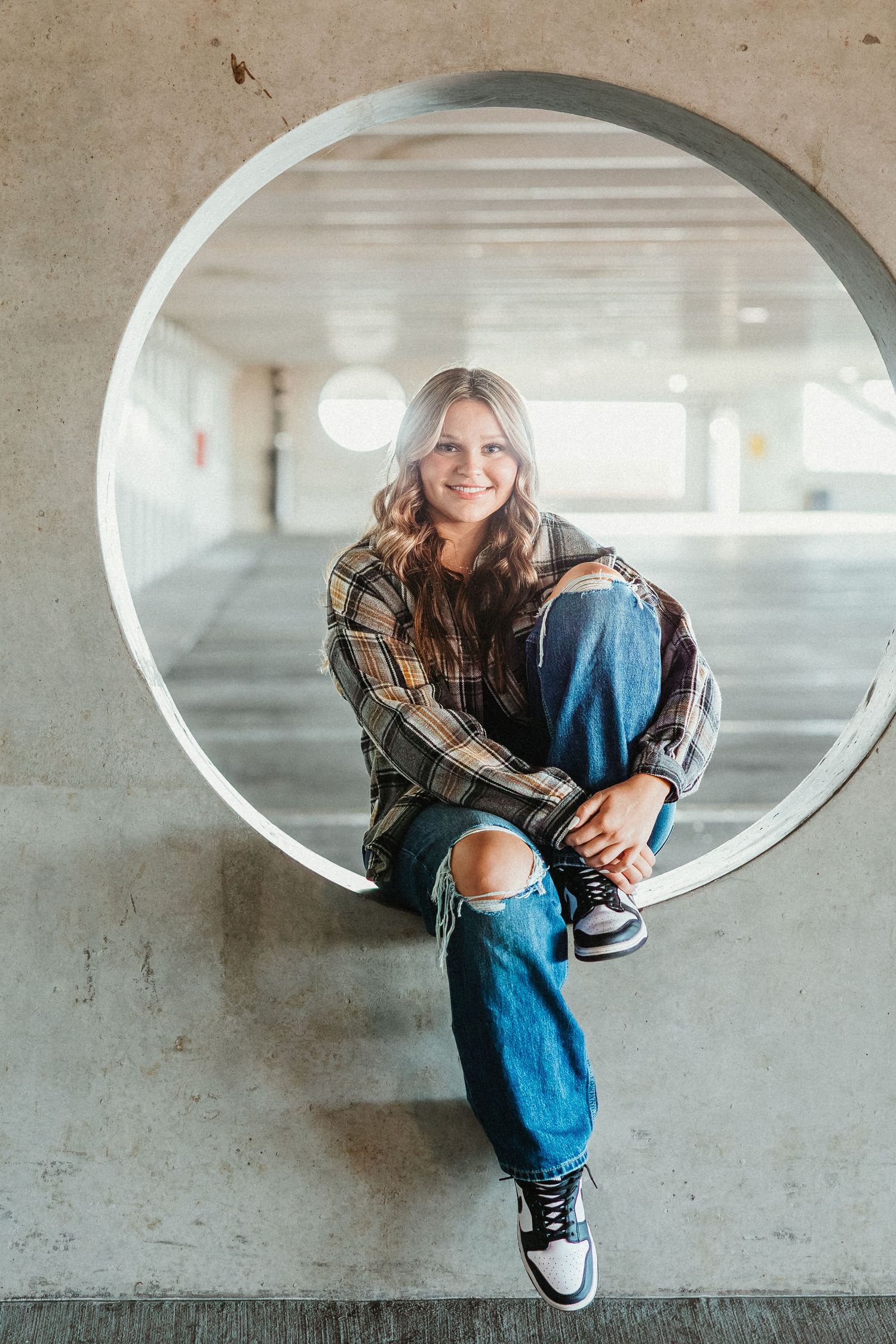 Casual portrait in plaid shirt and ripped jeans seated in circular concrete window.