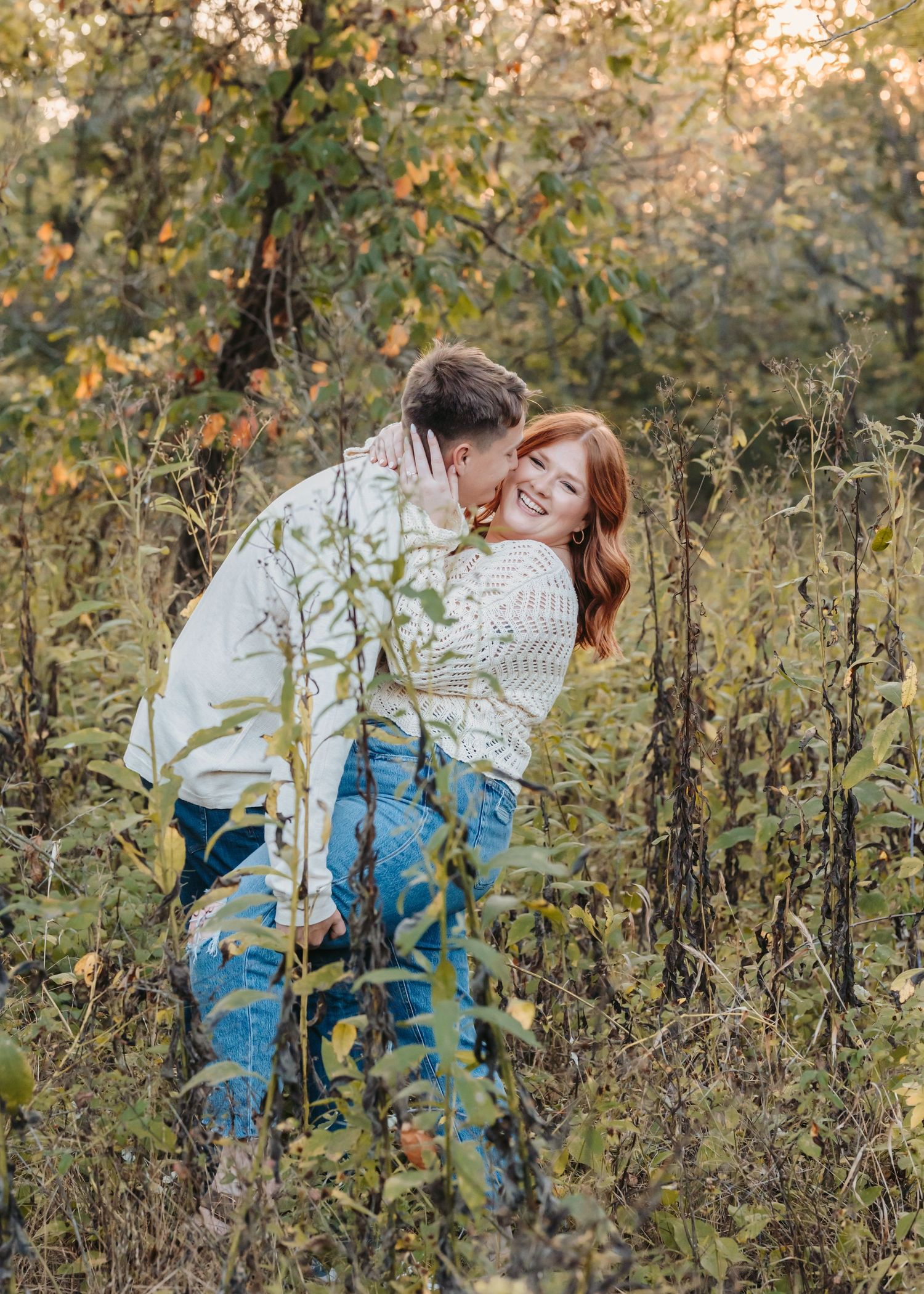 A romantic couple embraces while standing in tall grass during golden hour.