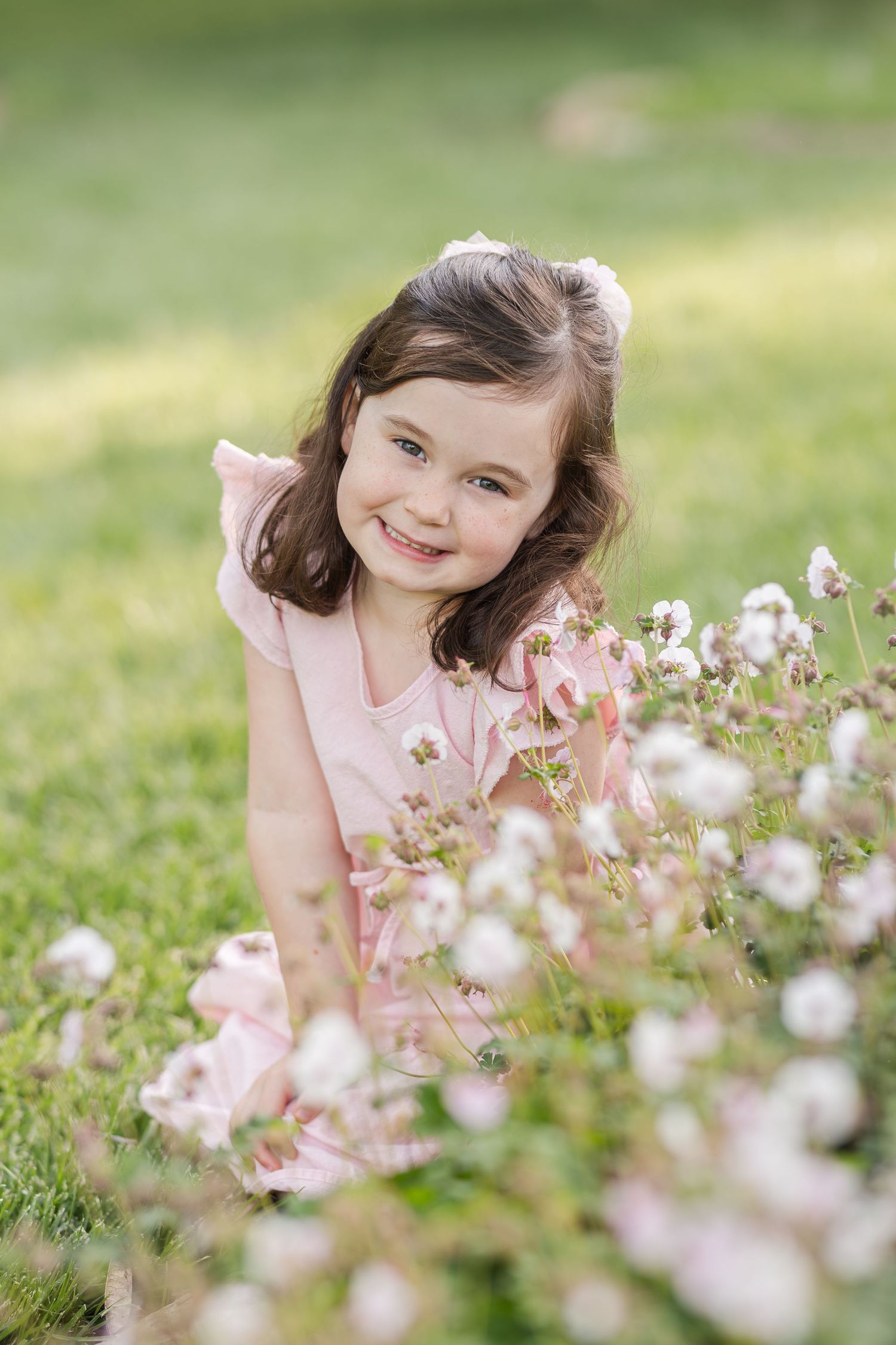 A young child in a pink dress sits among delicate white wildflowers in a sunlit meadow with a soft bokeh effect.