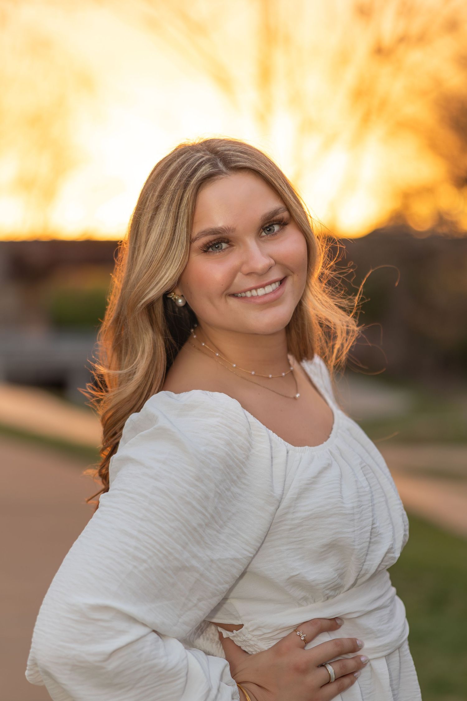 Portrait of a person in a white long-sleeved dress smiling warmly during golden hour sunset outdoors.
