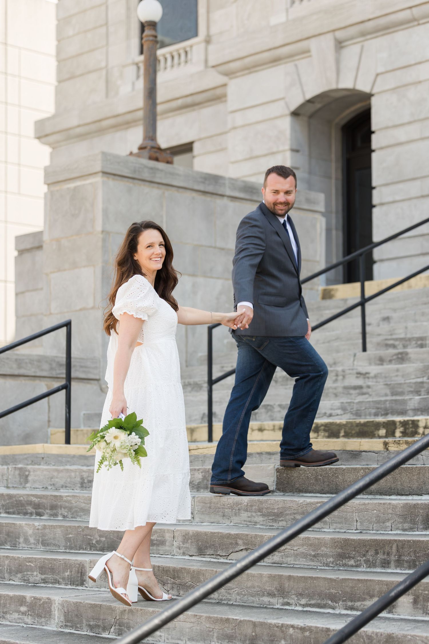 A couple in wedding attire descends stone steps outside a grand building with the bride holding a white floral bouquet.