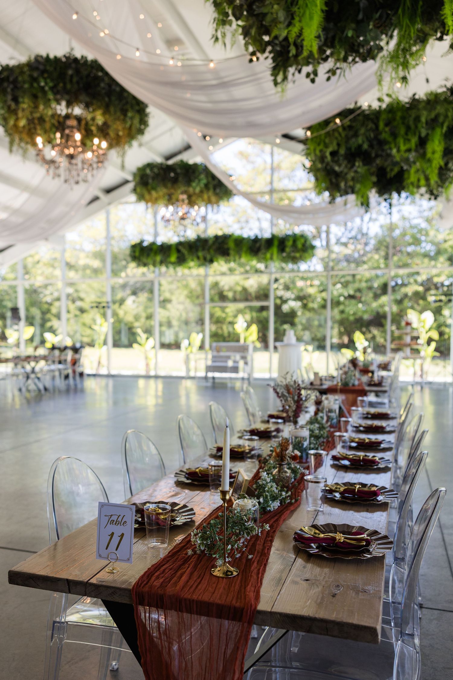 Elegant wedding reception table set in a greenhouse venue with greenery ceiling installations and ghost chairs.