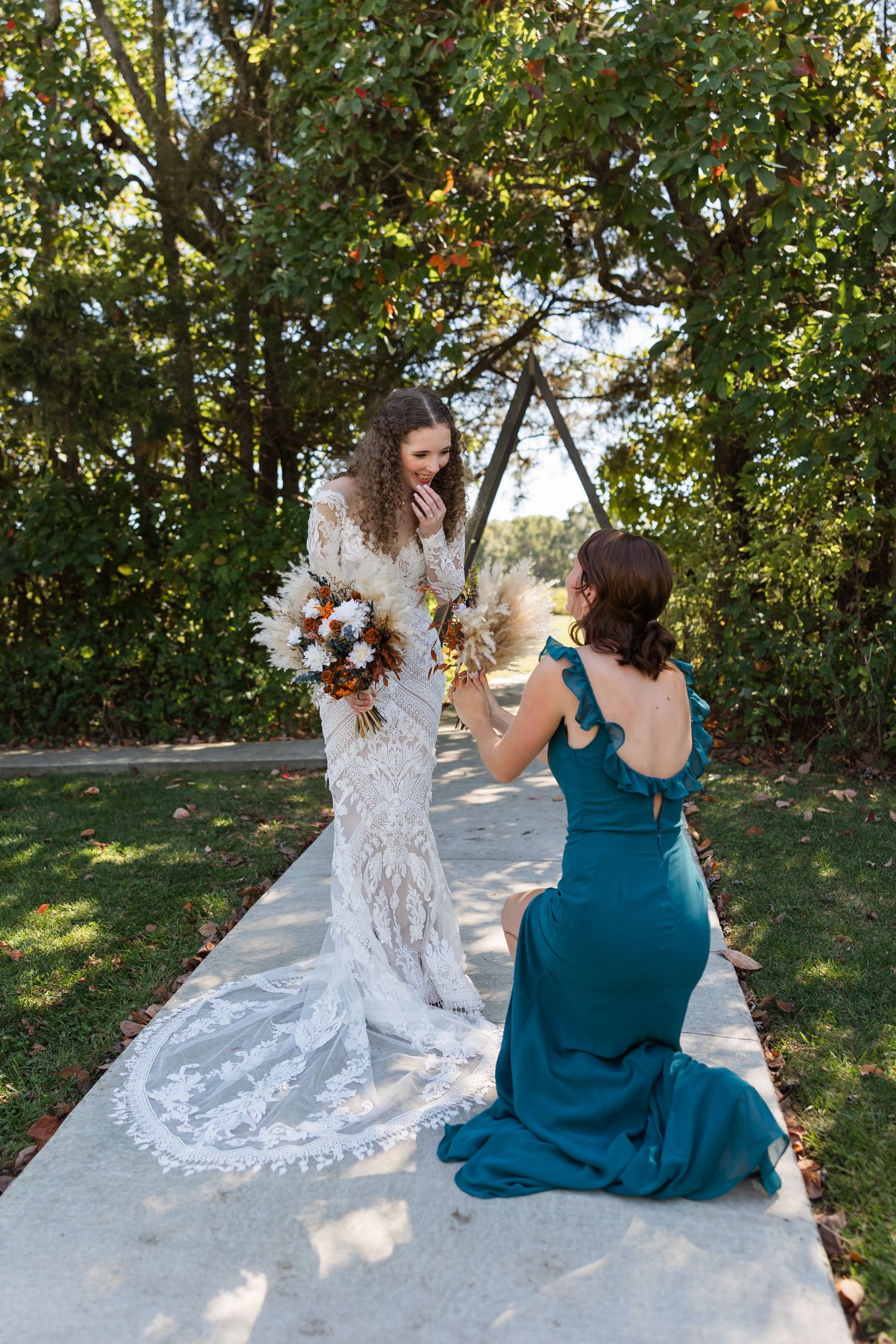 Bridesmaid in teal gown kneels to adjust bride's lace wedding dress train in garden setting.