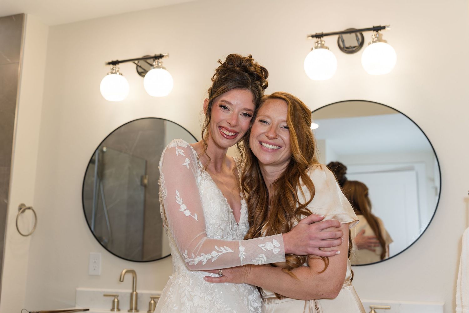 Two women embrace and smile in a bright bathroom, one wearing a lace bridal gown before her wedding day.