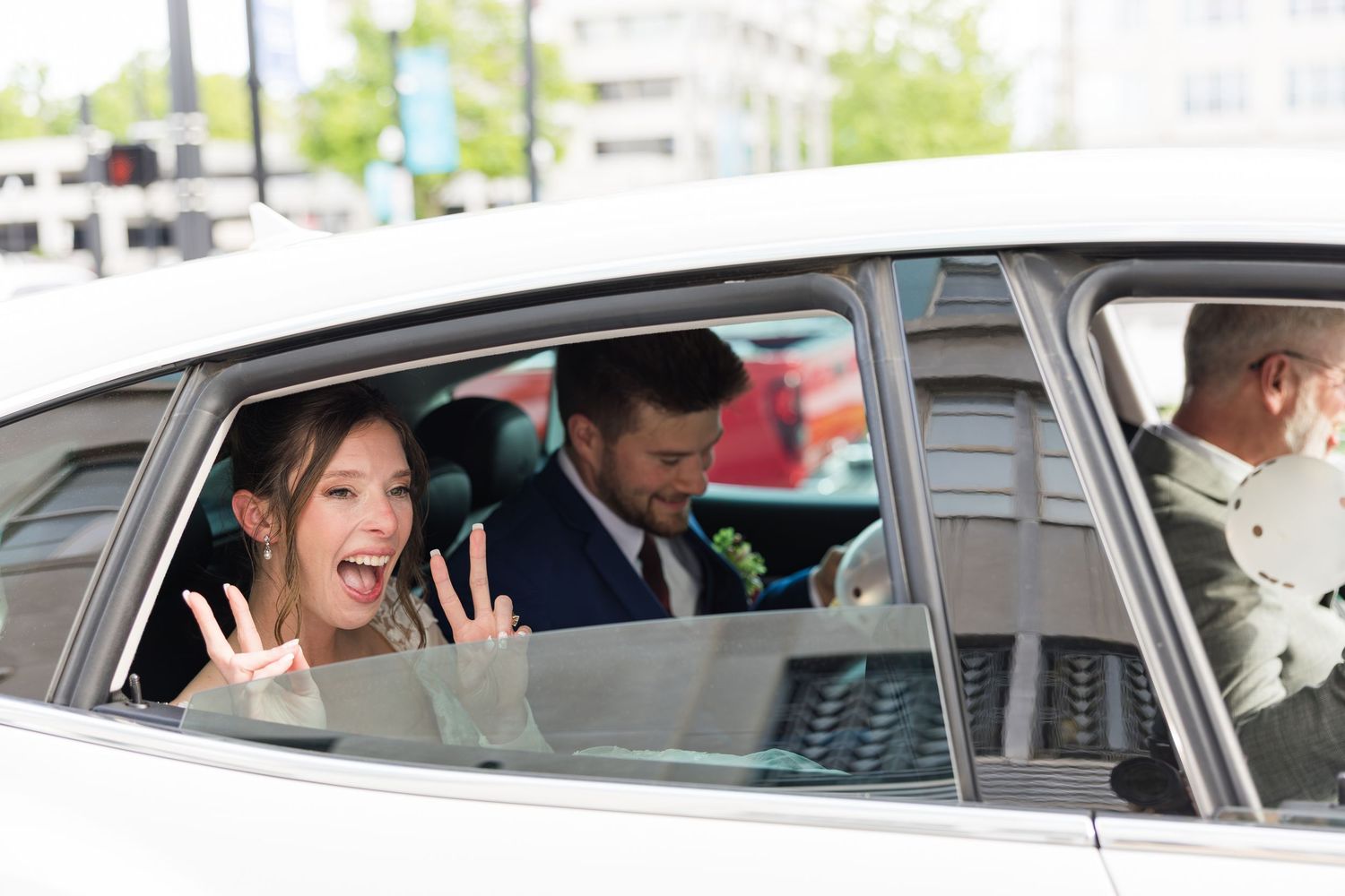 Excited bride waves from vintage white getaway car with groom after wedding ceremony.
