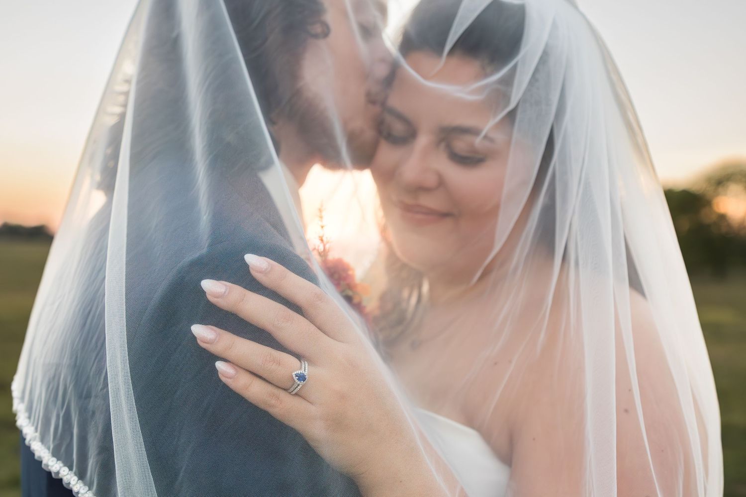 Bride and groom share intimate moment under wedding veil at golden hour sunset, showcasing elegant engagement ring.