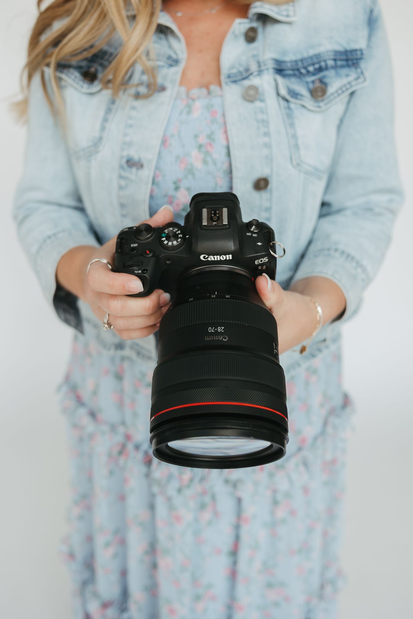 A person wearing a denim jacket holds a Canon DSLR camera against a white background.
