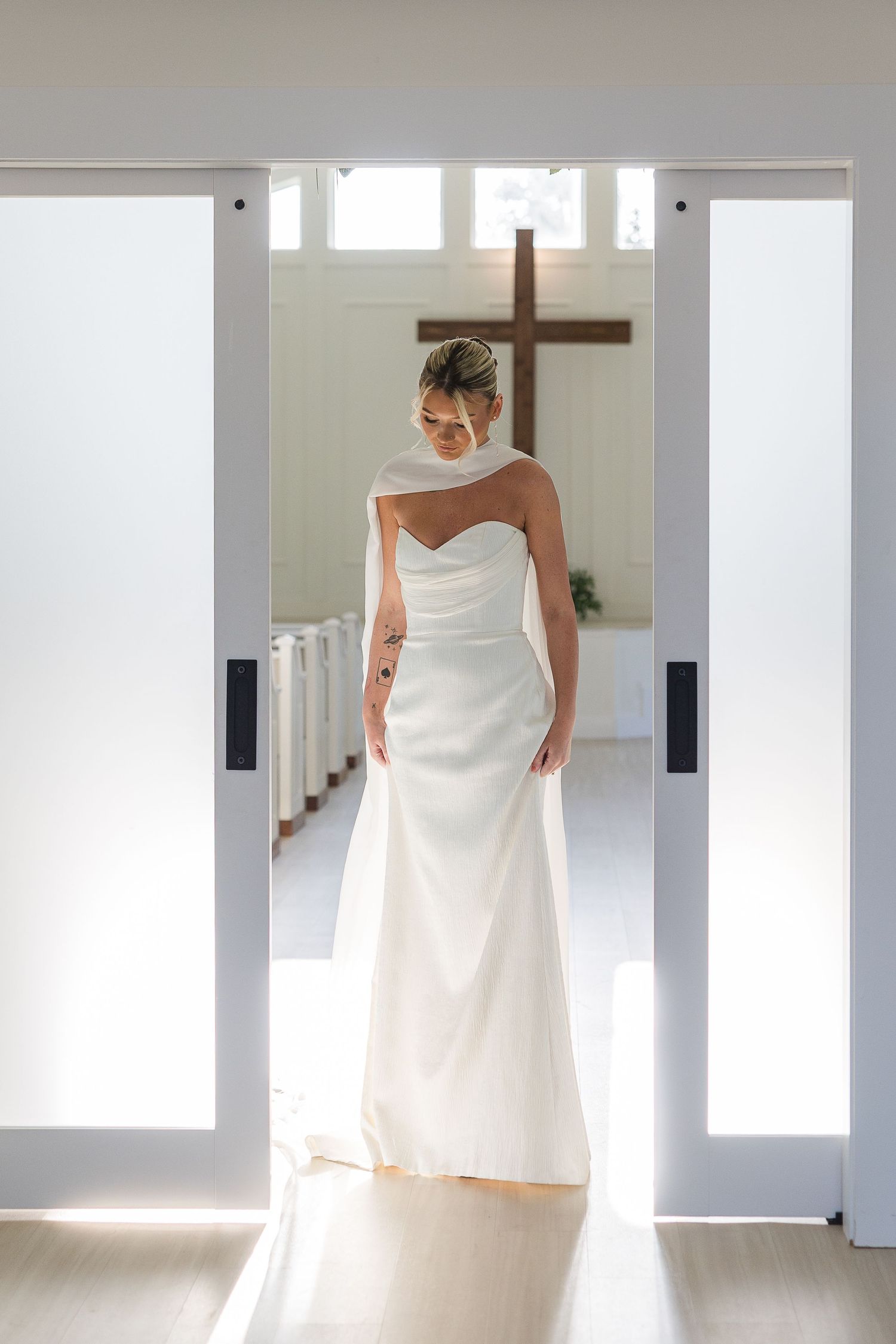 A bride in an elegant strapless white gown stands in a doorway with a wooden cross visible in the background.